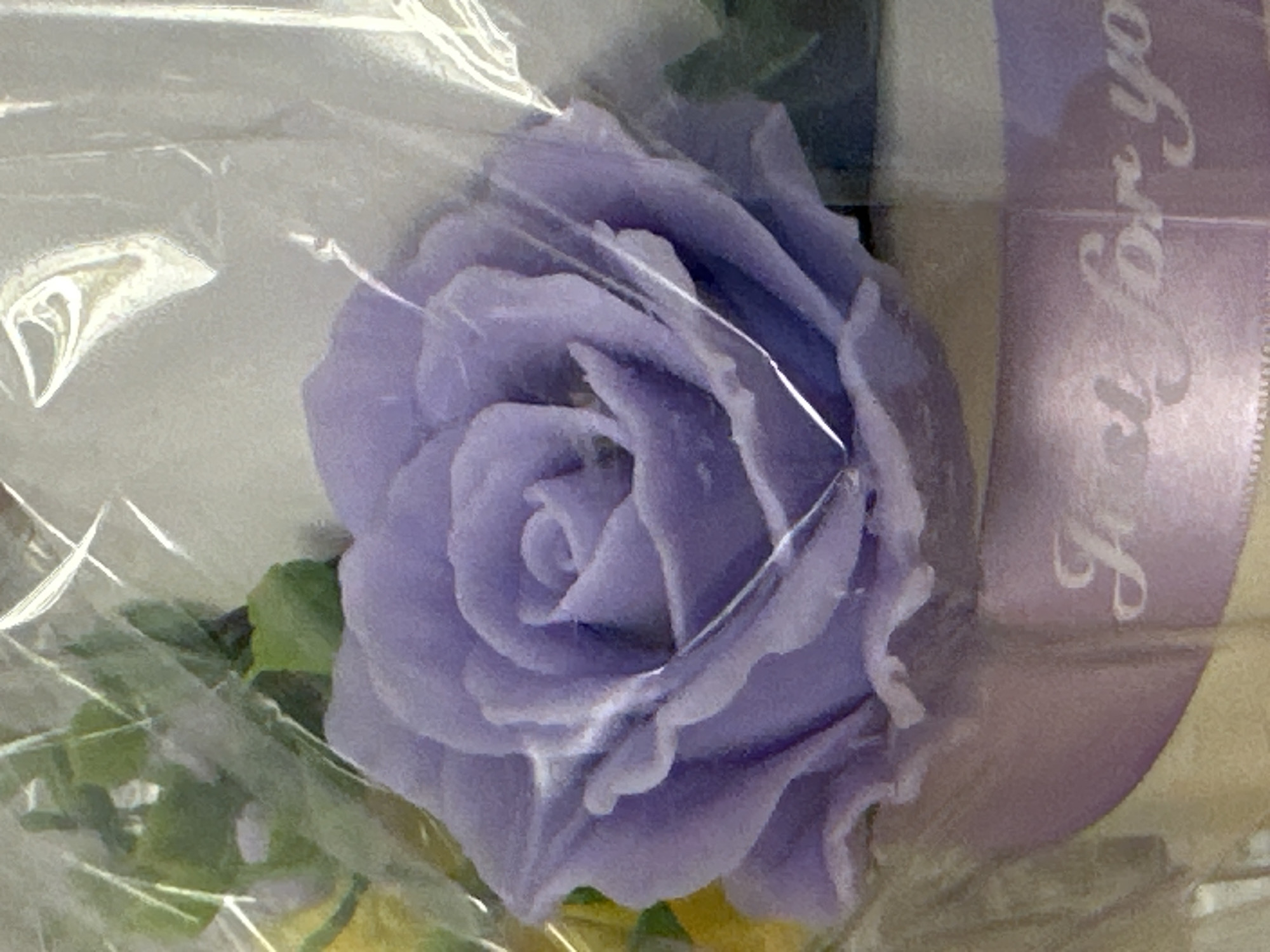 The image shows a close-up of a lavender rose-shaped object, possibly a soap or a candle, wrapped in clear plastic. The object has intricately detailed petals. A lavender ribbon with the embossed text "Just for you" is visible at the bottom. Green foliage and a hint of yellow are also present in the background. The overall impression is of a gift item. The image does not provide any information about the city of Yessentuki, Russia, nor does it contain any people or specific environmental details that could be used to determine a location.
