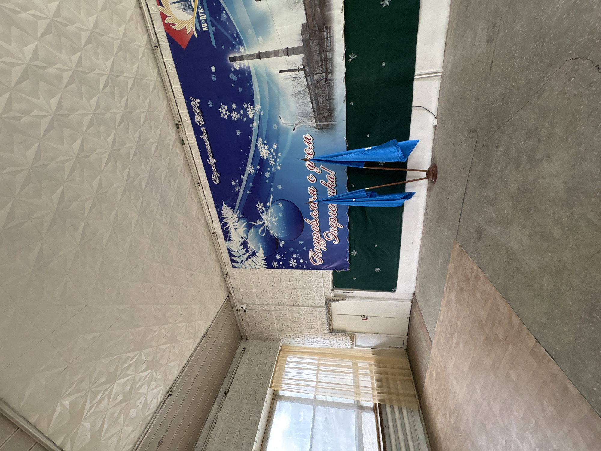 The image shows a room with a textured white ceiling and walls, and a floor with sections of light brown and gray tiling. There is a window with a sheer curtain at the bottom of the frame. Above the window, a banner hangs with a blue background decorated with snowflakes and a stylized industrial scene with a tall smokestack. The banner includes the text "Биробиджанская ТЭЦ", "Поздравляем с днем Энергетика!", and "АО «ДГК»". A stand with two small blue flags is in front of the banner. The context clues provided suggest that the location is in Birobidzhan, Russia.