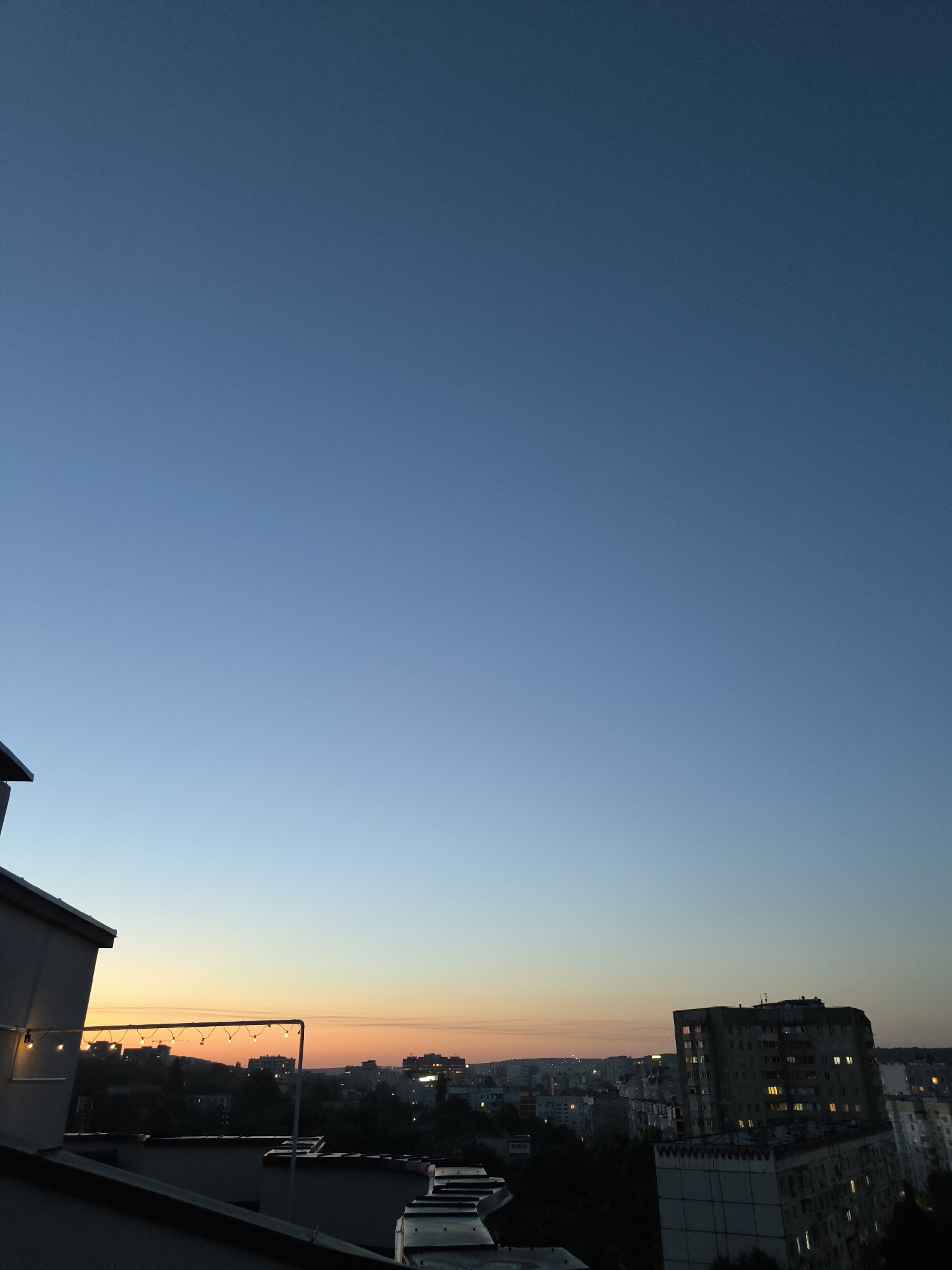 The image depicts a city skyline at dusk, likely in Chișinău, Moldova, as indicated by the architectural style of the buildings. The sky transitions from a deep blue at the top to a soft orange hue near the horizon. In the foreground, the edge of a rooftop is visible, with a string of lights draped across a railing. Beyond this, a cluster of residential buildings, characterized by their multi-story Soviet-era architecture, dominates the middle ground. Some of the apartment windows are illuminated, suggesting evening activity within. The overall atmosphere is tranquil, capturing the peaceful transition from day to night over an urban landscape.