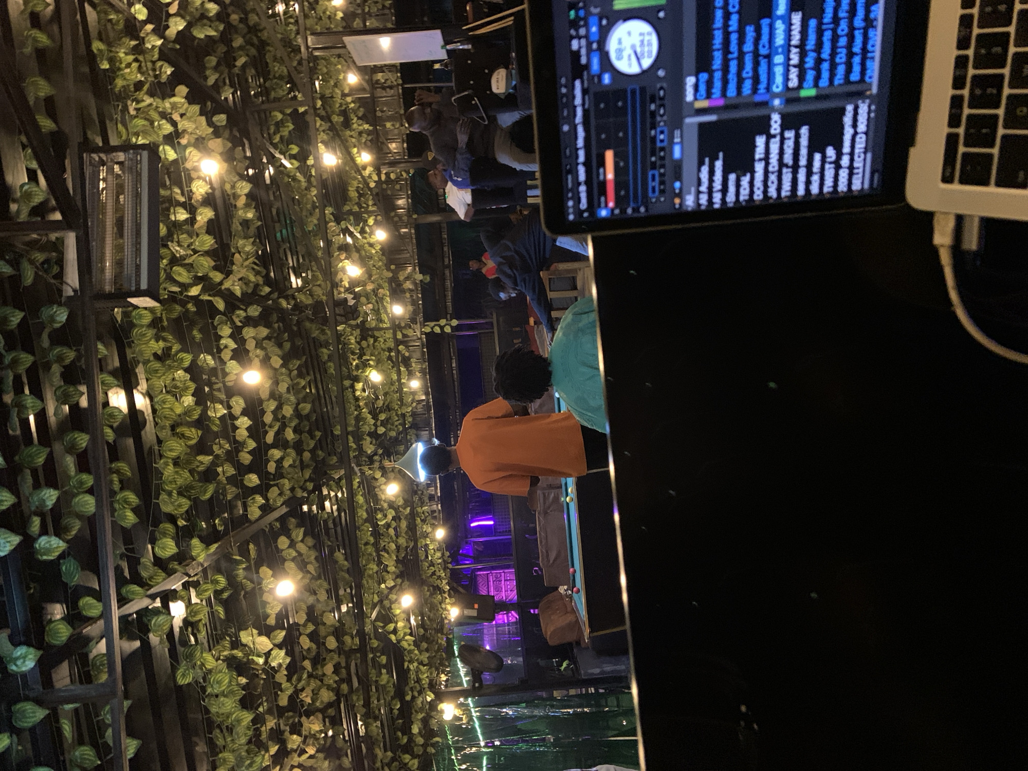 A low-angle, indoor shot captures a dimly lit, open-air venue adorned with artificial green vines and string lights on the ceiling. In the foreground, a laptop displays a digital audio workstation interface, suggesting a DJ setup. In the midground, a group of individuals are engaged in activities around a pool table. One person in an orange shirt stands near the pool table, while others are either playing or observing. Further in the background, more people are visible, some interacting and others in motion. The overall atmosphere appears to be a casual social gathering.