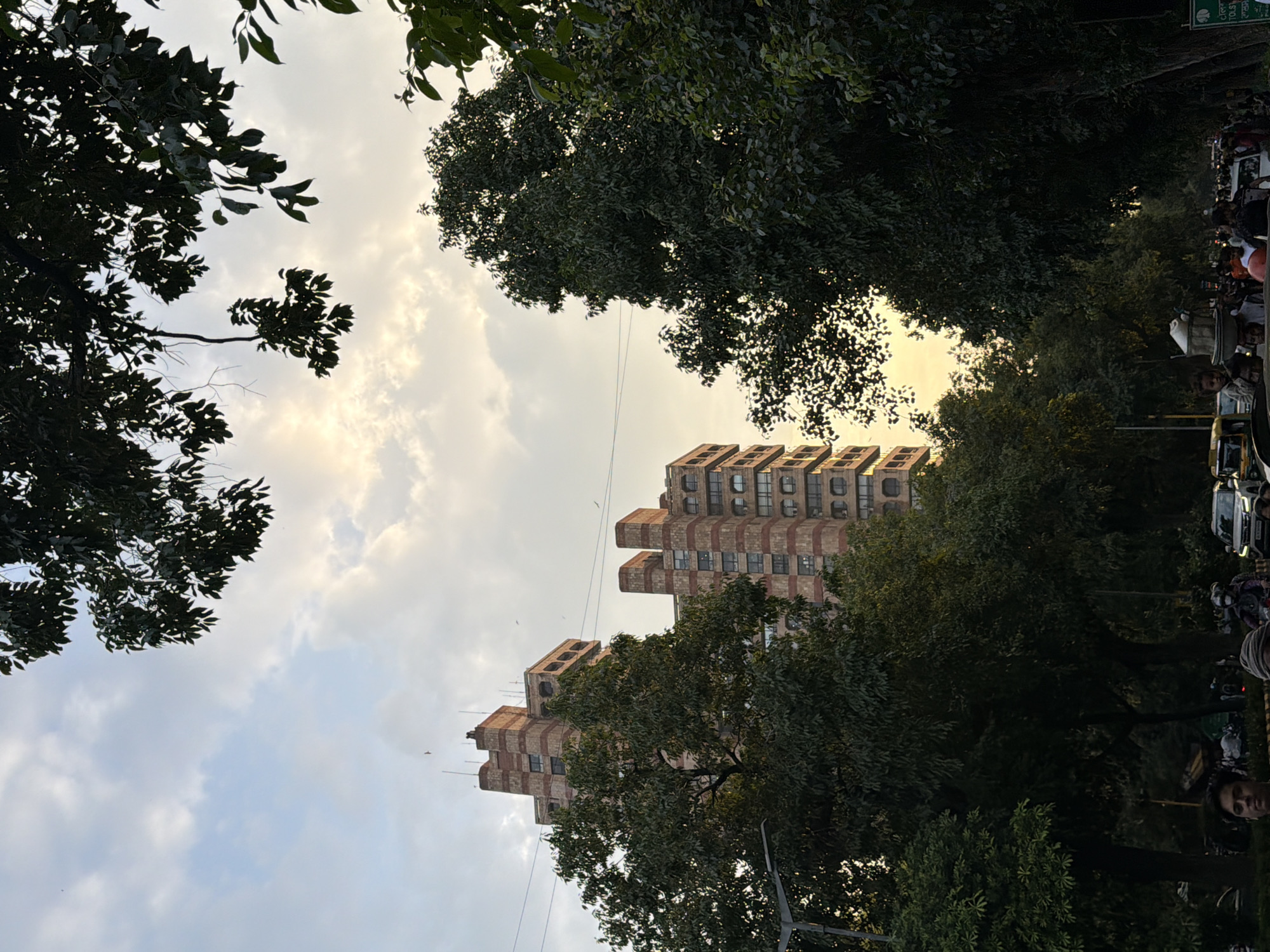 This is a photograph taken in New Delhi, India, likely in the late afternoon or early evening, given the lighting. The image captures a street scene with significant foliage, prominent architecture, and a cloudy sky.
In the background, two tall, rectangular buildings stand out. They are constructed with a distinctive pattern of reddish-brown bands alternating with rows of windows. These buildings appear to be modernist in design, with stepped sections and recessed balconies or openings.
In the foreground and midground, lush green trees with dense canopies dominate the scene, partially obscuring the buildings and creating a layered effect. The branches and leaves of these trees frame the sky at the top of the image. The sky itself is filled with clouds, with patches of sunlight breaking through, creating a soft, diffused illumination.
The street level appears to be bustling with activity. Several vehicles, including cars and what looks like an auto-rickshaw, are visible in traffic. There are also many people on the street, some walking and others on motorcycles or scooters. Some individuals are carrying items, such as a person with a large vessel on their head. The presence of a lamppost on the left side of the frame further indicates an urban street environment. The overall impression is one of a busy city street integrated with significant greenery, common in parts of New Delhi.