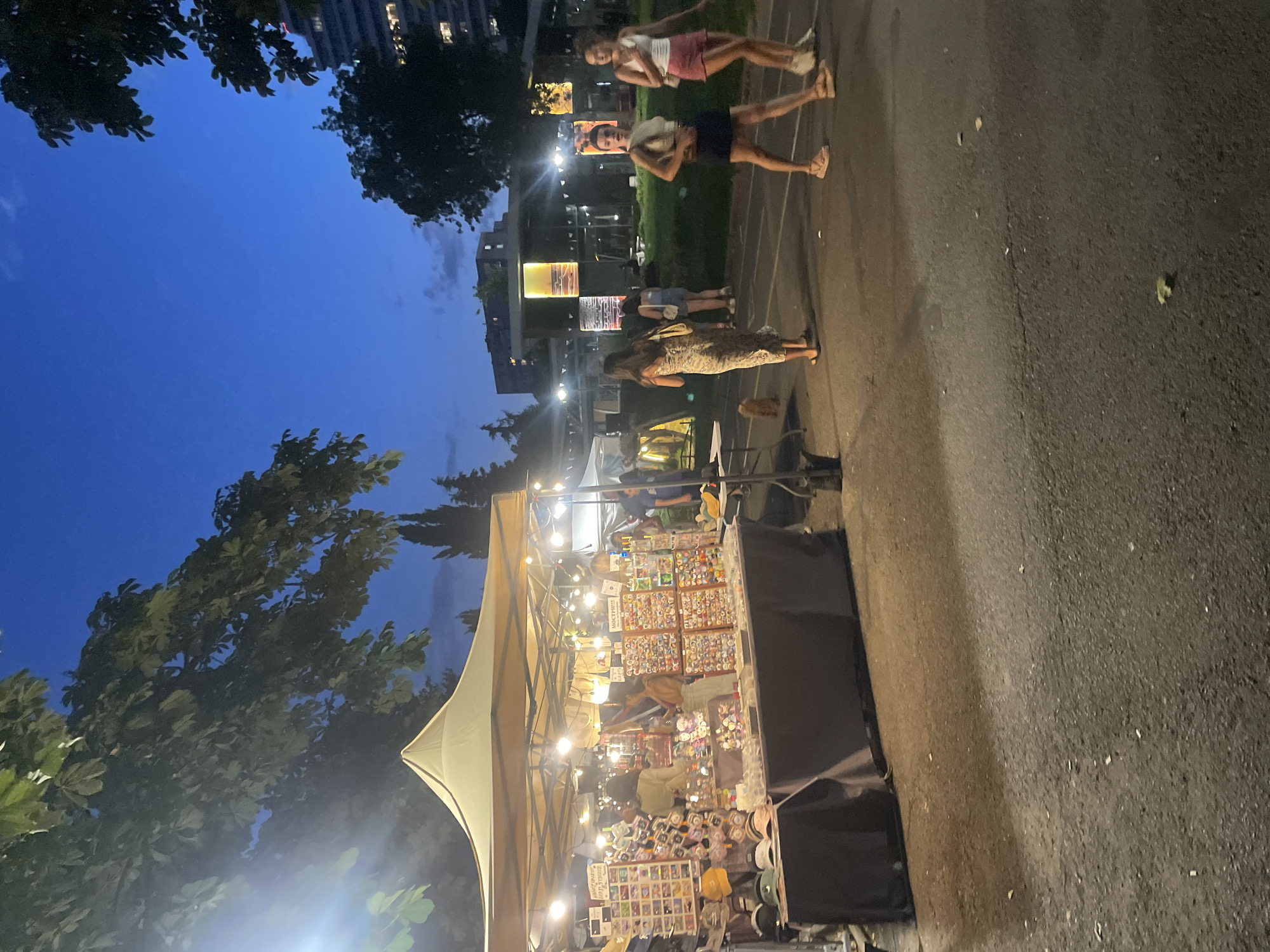 The image depicts an outdoor night market at the Sea Garden in Burgas, Bulgaria. A brightly lit stall, covered by a beige tent, is set up and displays a variety of goods, including hats, pins, and possibly jewelry. Several people are browsing the stall and walking in the vicinity. The sky is a deep blue, indicating it is twilight or early night. Trees and buildings are visible in the background, and string lights are hung between poles. The ground appears to be paved. The overall atmosphere is relaxed and communal, suggestive of a summer evening event.