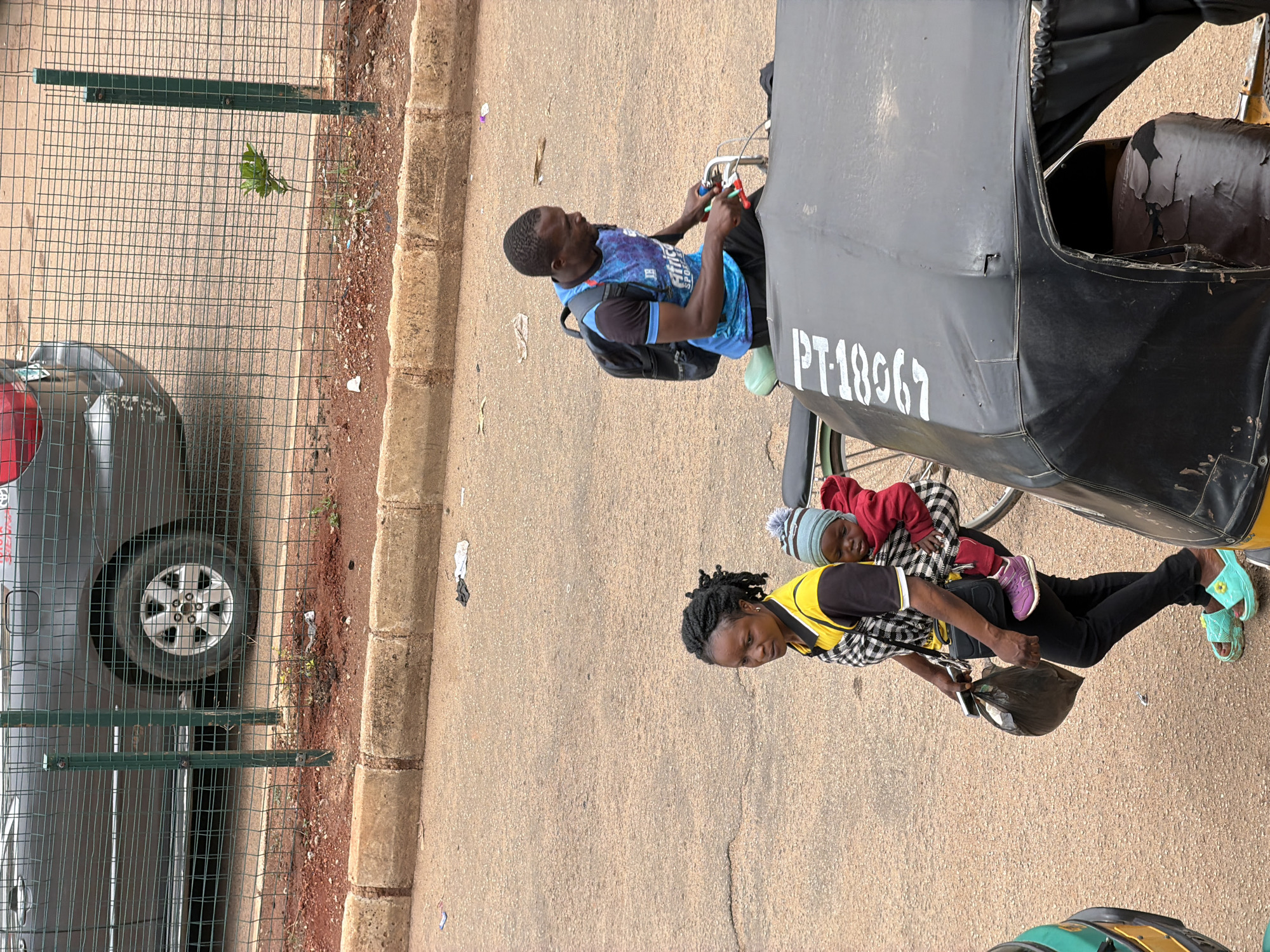 In Bukuru, Nigeria, on a dusty asphalt road, a woman with braided hair walks, carrying a toddler strapped to her back. She wears a yellow and black checkered top, black pants, and turquoise sandals. A black plastic bag hangs from her left hand. Beside her, a man on a bicycle, wearing a blue and black t-shirt and a backpack, rides past. The bicycle has a black seat and a green frame. A three-wheeled auto-rickshaw, with a black canopy bearing the white markings "PT-1867", is parked on the right side of the frame. Behind the people and vehicles, a dark-colored car is partially visible behind a green metal fence, with its front wheel and part of the body showing. The ground near the fence is covered in dirt and some scattered debris.