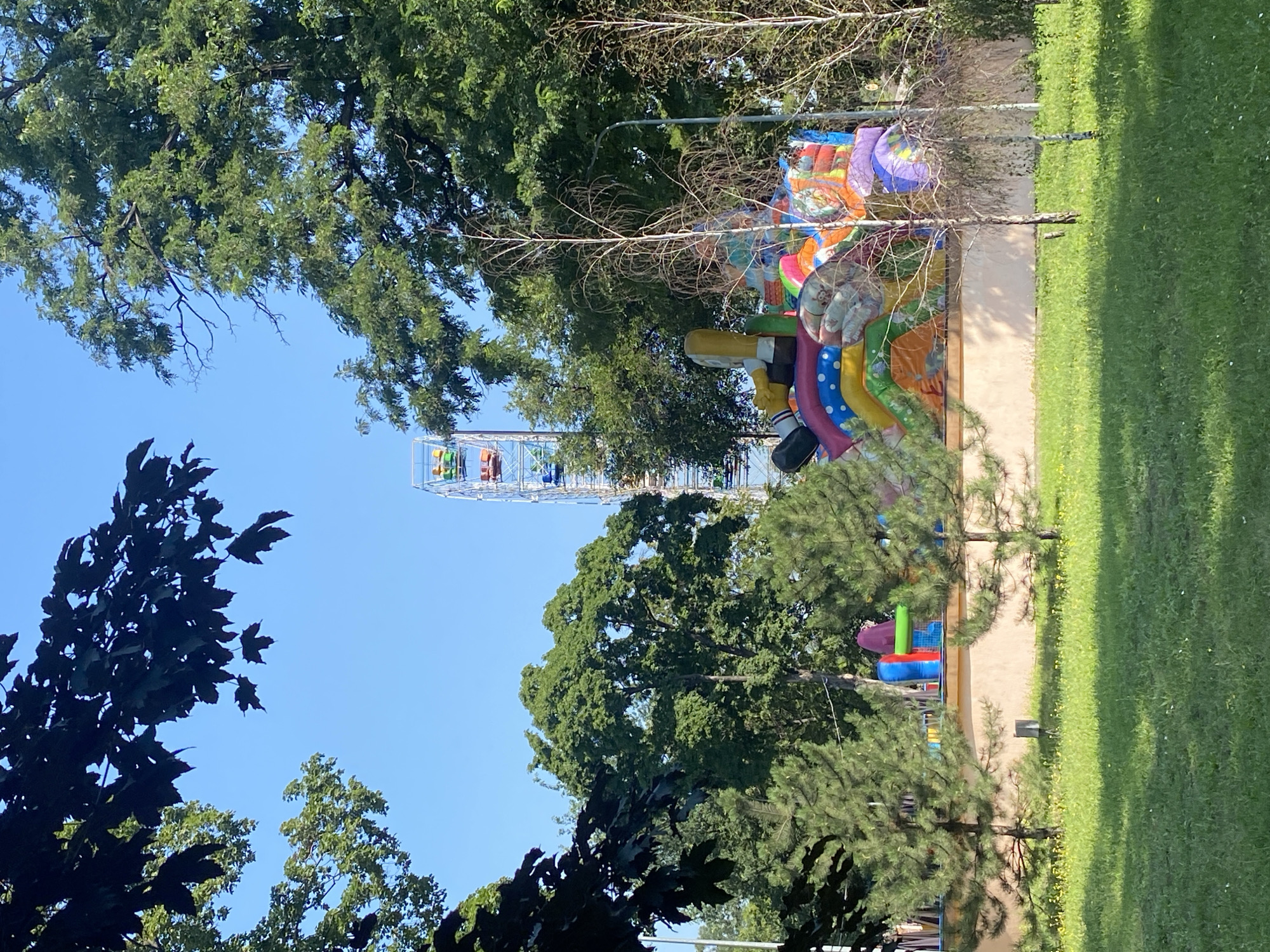 In Kamianske, Ukraine, a vibrant amusement area is depicted under a clear blue sky. The scene is framed by the dark green foliage of mature trees in the foreground, with lighter green trees and a bright blue sky visible beyond. Dominating the mid-ground is a large, colorful inflatable playground with multiple sections, featuring patterns of stripes, polka dots, and cartoonish designs. Partially visible within the inflatable structure is a figure resembling Homer Simpson, dressed in his signature white shirt and blue pants, with yellow skin. To the left of the inflatable, a tall, white Ferris wheel stands with several colorful gondolas. The Ferris wheel is partially obscured by trees. In the foreground, a well-maintained green lawn is dotted with small yellow wildflowers. The background consists of a light-colored wall, likely part of the amusement area's boundary, and more trees. The overall impression is one of a sunny day at an outdoor entertainment venue.