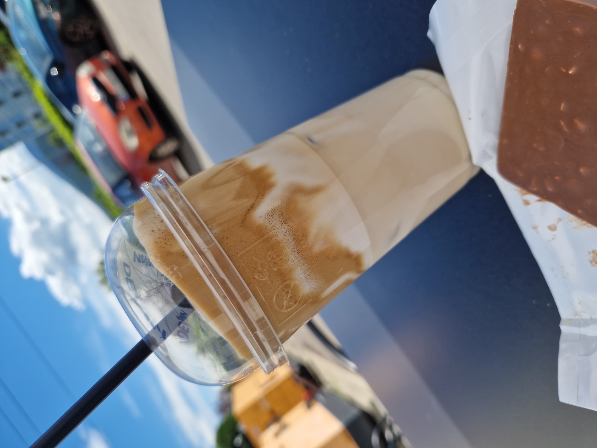 A close-up, outdoor shot features a plastic cup filled with a layered beverage, likely Dalgona coffee, with a black straw inserted. The drink has a foamy, light brown top layer over a milky white liquid. The cup is positioned on a dark, glossy surface, possibly a table, with a piece of chocolate bar, wrapped in white paper, visible in the foreground. In the background, a clear blue sky with white clouds suggests a sunny day. Out of focus, there are cars parked, including an orange vehicle and a darker one, along with a building and yellow boxes. The general setting appears to be an urban area, with the presence of cars and buildings hinting at a street or parking lot. The overall composition is angled, with the coffee cup prominently displayed. There are no visible people in the image. The location is identified as Sofia, Bulgaria.