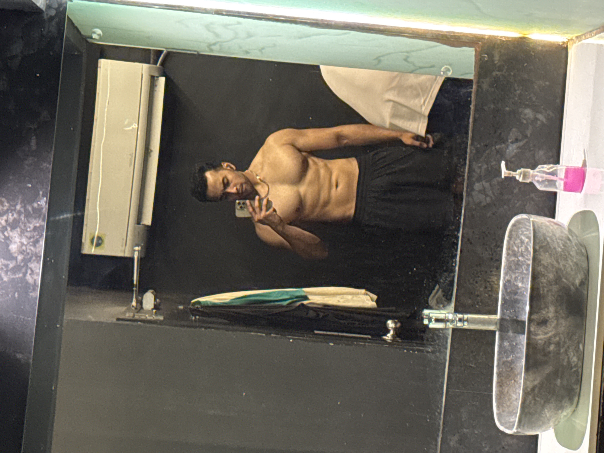 A shirtless man with dark hair and a gold chain is taking a mirror selfie in a bathroom with black walls. He is holding a smartphone and wearing black pants. The bathroom has a white hand towel hanging, a white hand dryer, a partially visible green and white towel, a grey sink, and a pink soap dispenser. The location cannot be definitively determined to be Kolkata, India.