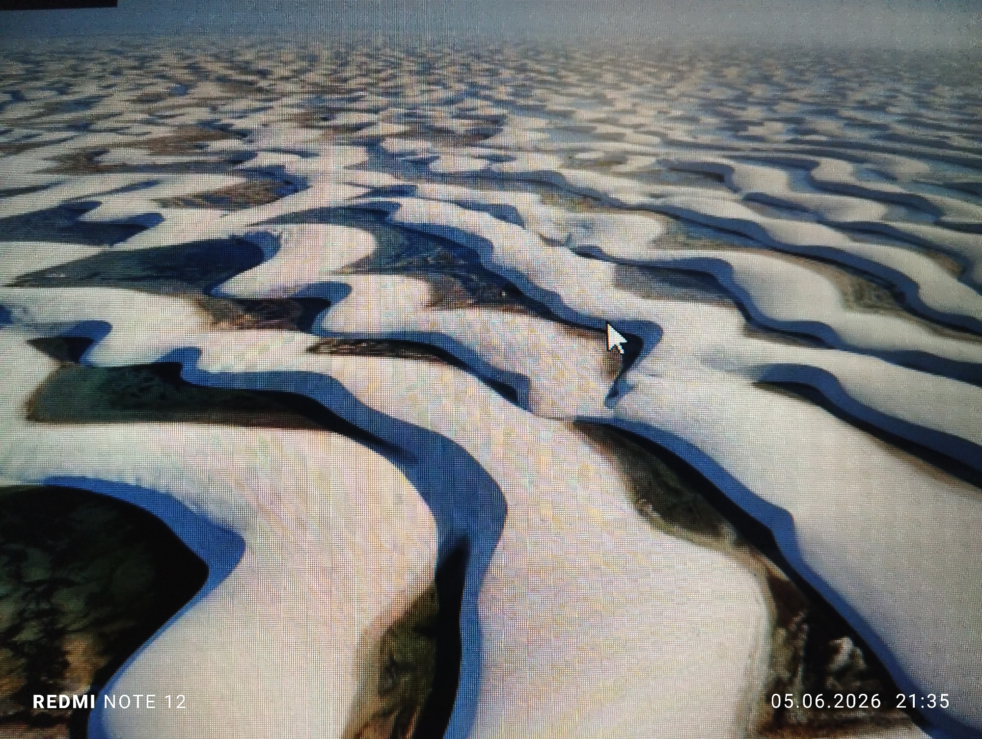 The image displays a high-angle view of a natural landscape, which is being viewed on a digital screen, as indicated by the visible pixel grid and the presence of a white, angled mouse cursor in the mid-right portion of the frame. The landscape is characterized by numerous curvilinear, light-colored (predominantly white) sand formations resembling dunes, intricately intertwined with dark blue and dark green water bodies or lagoons. These elements form a complex, sprawling pattern extending towards a hazy horizon. Shadows cast by the sand formations suggest a distinct light source. There are no individuals, animals, or man-made structures visible within the depicted natural environment. Overlaid text in the bottom left corner reads "REDMI NOTE 12", and in the bottom right, "05.06.2026 21:35". The scene is located in the city of Burgas, Bulgaria.