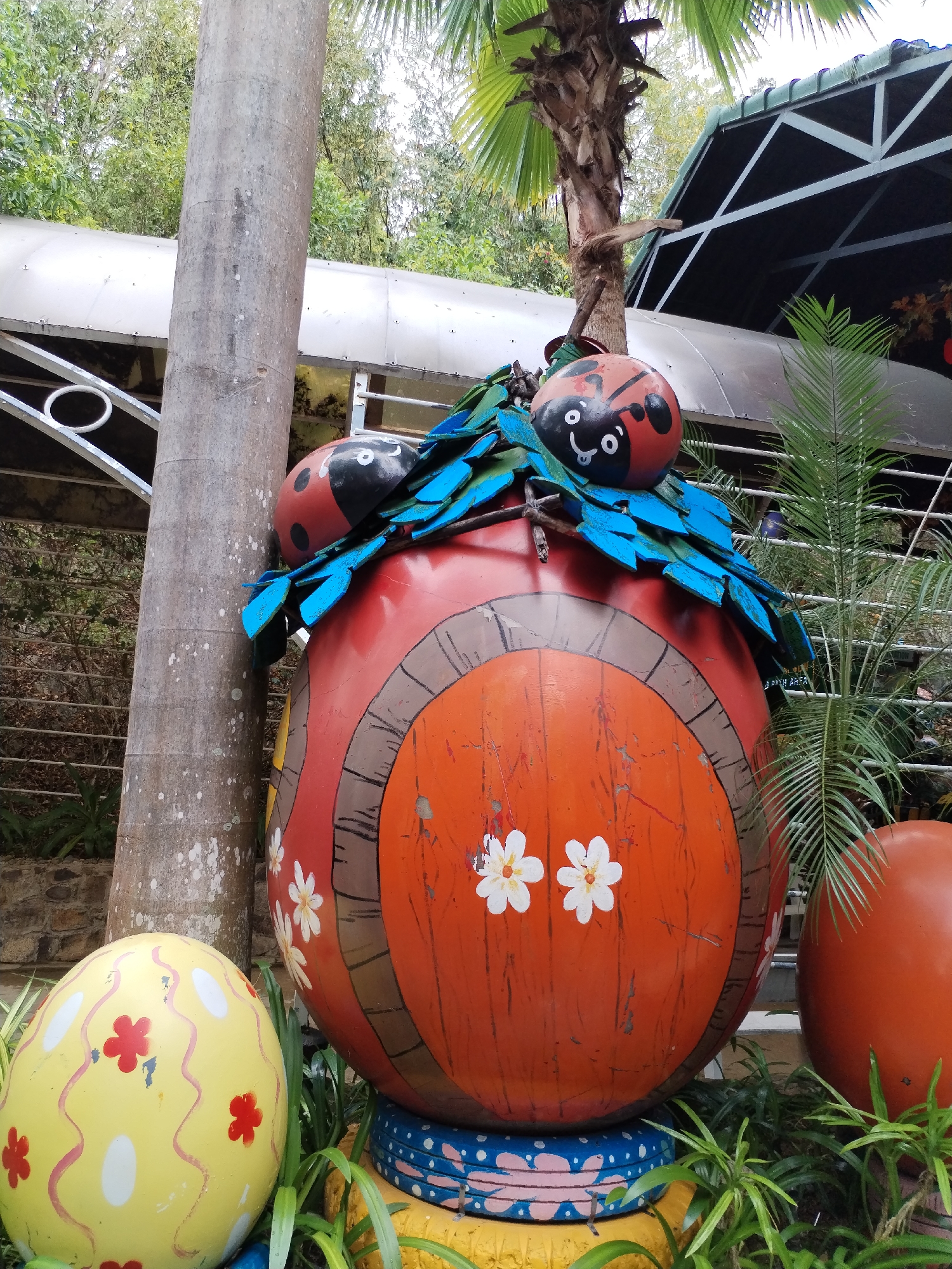 Captured in Nam Nha Trang Ward, Vietnam, the scene showcases a whimsical outdoor decoration. A large, egg-shaped structure painted red and orange resembles a miniature house, complete with a door decorated with white flower motifs and a roof adorned with blue painted leaves and two ladybug figures. The "house" sits atop a stack of colorful painted tires surrounded by greenery. To its left is a yellow painted egg with red flower decorations. A cylindrical, mottled tree trunk stands beside the egg house. In the background, a partially visible structure with a metal frame and roof is present, along with a palm tree and other plants. The lighting suggests an outdoor setting during the day.