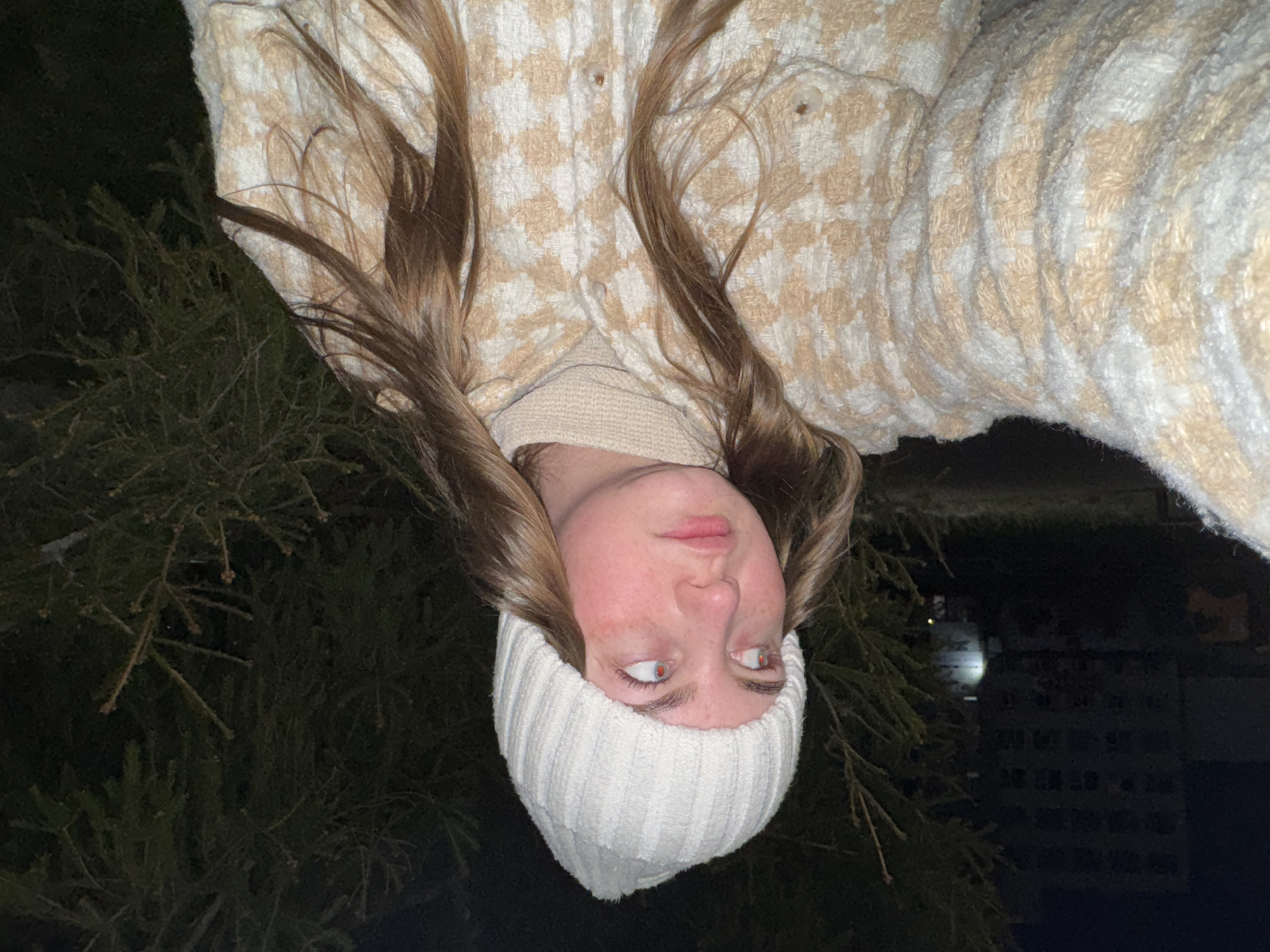A young woman is wearing a white beanie and a cream-colored houndstooth jacket. She is standing outside at night near a pine tree. In the background, there is a building that may be located in the city of Chaikovsky, Russia.