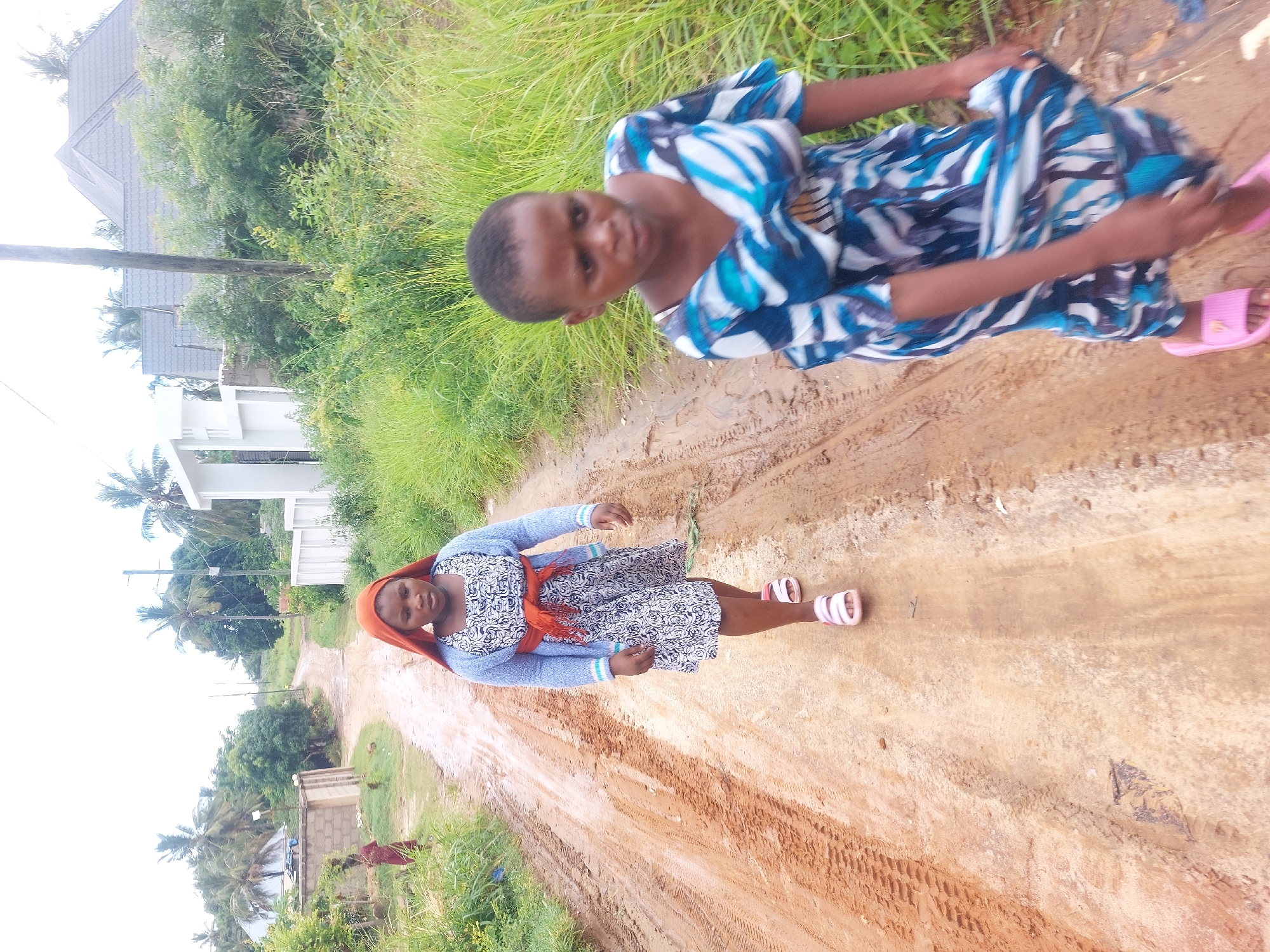 The image displays two young women standing on a muddy, unpaved dirt road, set in a residential or semi-rural area, consistent with parts of Dar es-Salaam, Tanzania. The photograph is oriented vertically.
In the foreground, towards the right, a woman with short dark hair wears a blue, black, and white patterned short-sleeved dress. She is looking towards the camera with a neutral to slightly smiling expression and is wearing pink slide sandals. Her right arm is visible, slightly extended.
Further back, towards the left, another woman is depicted. She is dressed in a light blue cardigan over a white and black patterned dress, with an orange headscarf or shawl draped over her head and neck. She also faces the camera, appearing calm with a gentle smile, and wears white and pink open-toed sandals.
The wide dirt road features distinct tire tracks and patches of mud, suggesting recent rainfall. Abundant green grass and bushes grow along both edges of the road. In the background, several houses with grey tiled roofs and light-colored walls are visible, interspersed with trees, including prominent palm trees. A wooden utility pole stands on the left side of the road. The sky appears bright and overcast. Both women seem to be casually posing for the photograph. No text is visible within the image.
