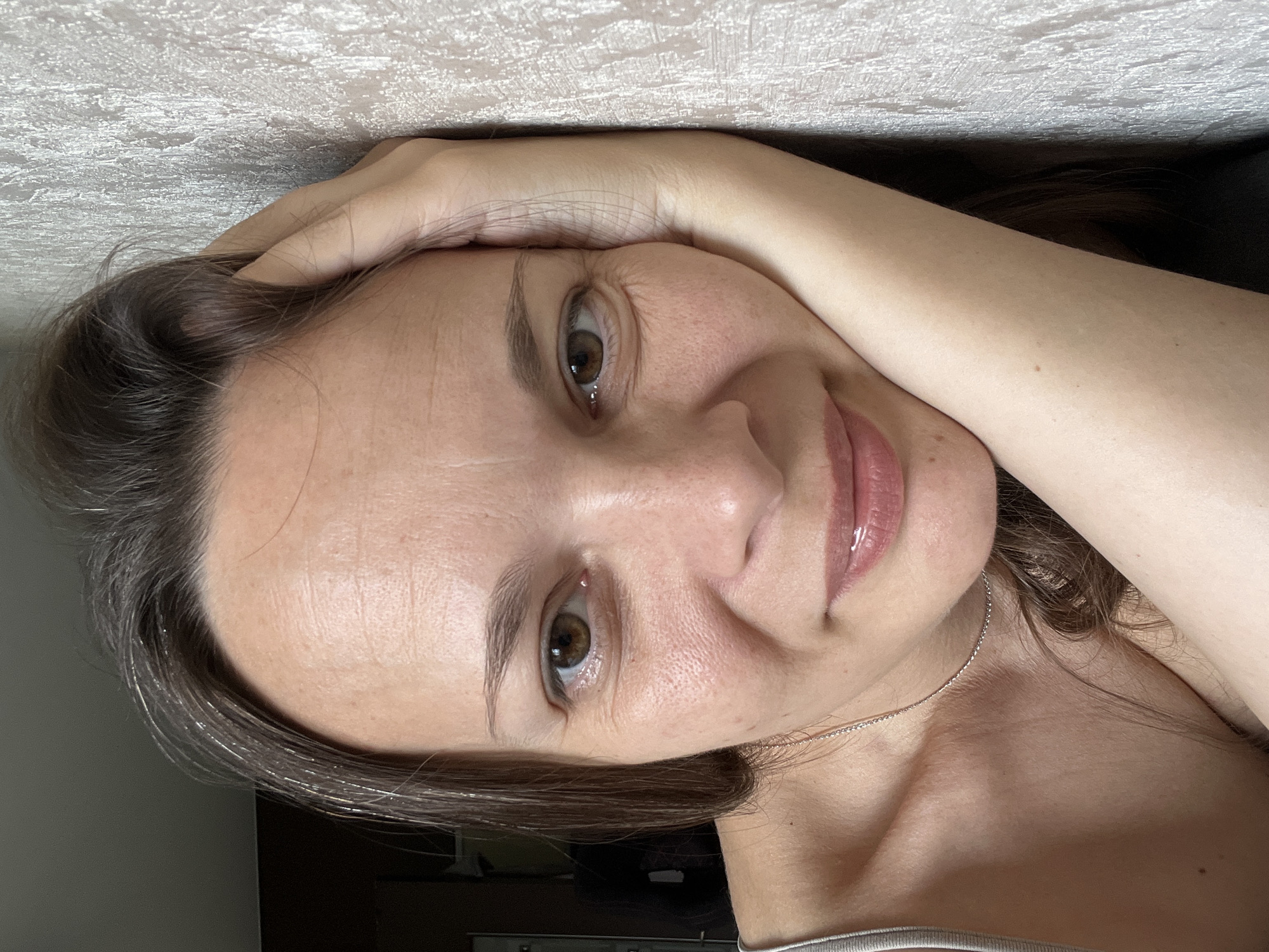 The image shows a close-up, head-and-shoulders portrait of a woman with brown hair and brown eyes. She is leaning her head on her hand, with her arm resting against a textured, light-colored wall. She is looking directly at the camera with a slight smile. She is wearing a light-colored tank top and a thin silver necklace. The background shows a dark area to the left and the textured wall to the right. The lighting suggests an indoor setting. There is no identifiable information to place the image in Tolyatti, Russia.