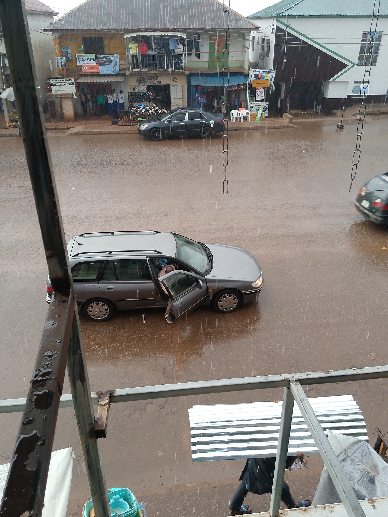 The image depicts a rainy day in Bukuru, Nigeria. A silver station wagon is parked on a waterlogged street with its driver's side door open. A person appears to be exiting the car. Across the street, there are buildings with shops displaying clothes and other merchandise. There are also parked cars. A metal structure with bars and some corrugated metal sheets is visible in the foreground, suggesting that the photograph was taken from an elevated position, possibly a balcony. Rain is heavily falling, creating puddles on the road.
