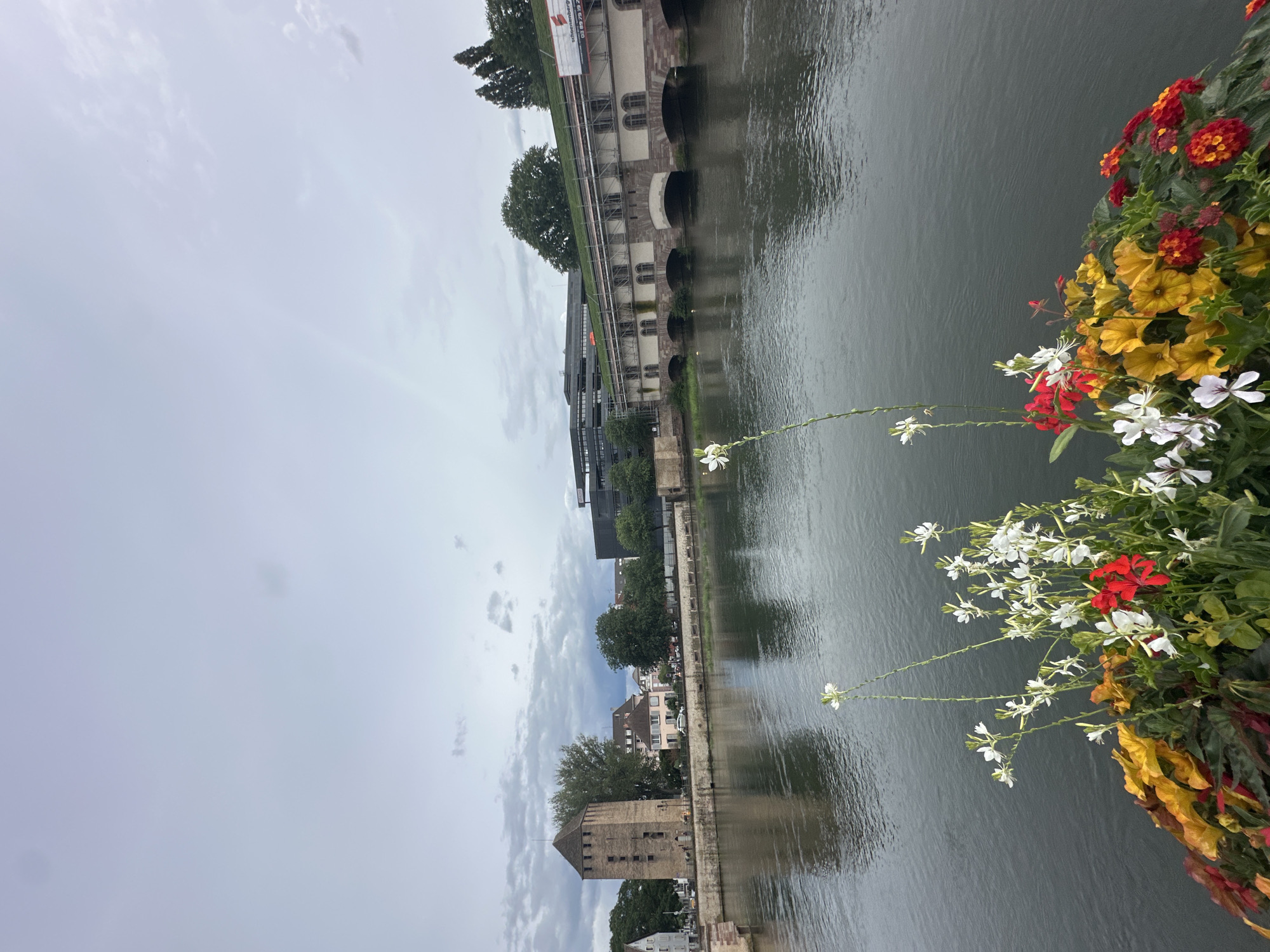The image shows a river in Strasbourg, France, with the historic Vauban Dam and fortifications visible in the background. A bridge with multiple arches spans the river, and a tall, brick tower, likely part of the city's ancient defenses, stands on the left bank. The sky is overcast, with a faint rainbow visible. In the foreground, a planter box overflows with a vibrant arrangement of red, yellow, and white flowers, adding a touch of color to the scene. The water of the river reflects the cloudy sky and the surrounding architecture.