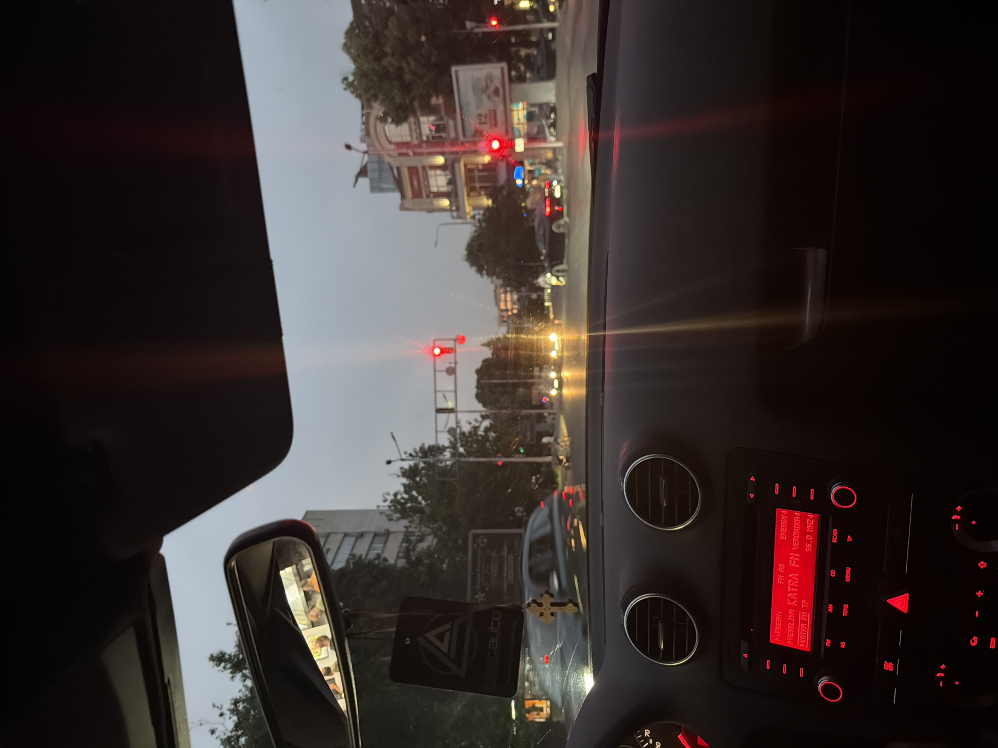 The image is taken from inside a car looking forward at a city street in Plovdiv, Bulgaria. The time appears to be dusk, with the sky a pale gray and streetlights beginning to illuminate the scene. Traffic lights are red, indicating the car is stopped.
In the foreground, the car's dashboard is visible, with a red-lit radio displaying "FRESH: FM AS ENERGY!", and "NEBELINA KATRA FM VERONIKA M 95.0 MHZI". Two circular air vents are above the radio. A dangling air freshener with a logo is hanging from the rearview mirror, which reflects people inside the car. A wooden cross is also hanging from the mirror.
The street outside shows several cars, including a dark colored sedan in the middle lane and a motorcycle in the distance. Buildings with illuminated windows line the street on the right side, and trees are visible. The traffic lights are red and prominently displayed at an intersection. The overall atmosphere suggests a typical urban evening commute or wait at a traffic signal.