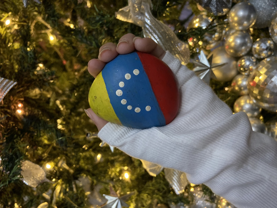 A hand in a white sweater holds a spherical object resembling a Christmas ornament painted with the Venezuelan flag. The ornament is divided into yellow, blue with white stars, and red sections. The background is a blurred Christmas tree decorated with lights and silver ornaments, creating a festive and celebratory atmosphere.