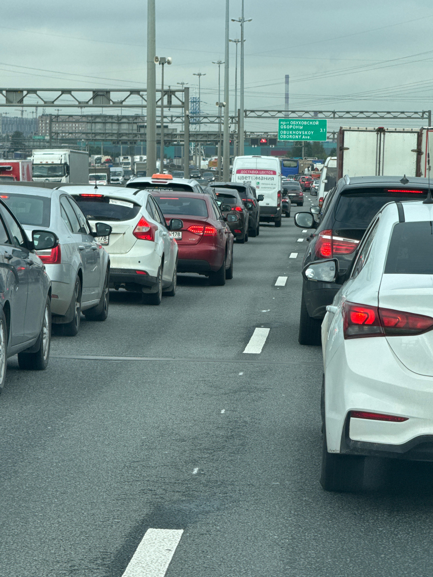 The image depicts a traffic jam on a multi-lane highway in Saint Petersburg, Russia. The sky is overcast, suggesting a cloudy day. In the foreground, several passenger cars are visible, including a silver sedan, a white hatchback, a red sedan, and a white sedan. Behind these, a line of trucks and vans continues, indicating a significant volume of traffic. Overhead, a green road sign with text in both Russian and English ("пр-т ОБУХОВСКОЙ ОБОРОНЫ", "OBUKHOVSKOY OBORONY Ave.") clearly marks the location as Obukhovskoy Oborony Avenue. The infrastructure includes overhead gantries, traffic lights, and numerous power lines, characteristic of a major urban transport artery. The overall scene conveys a standstill or very slow-moving traffic condition, a common occurrence on such routes.