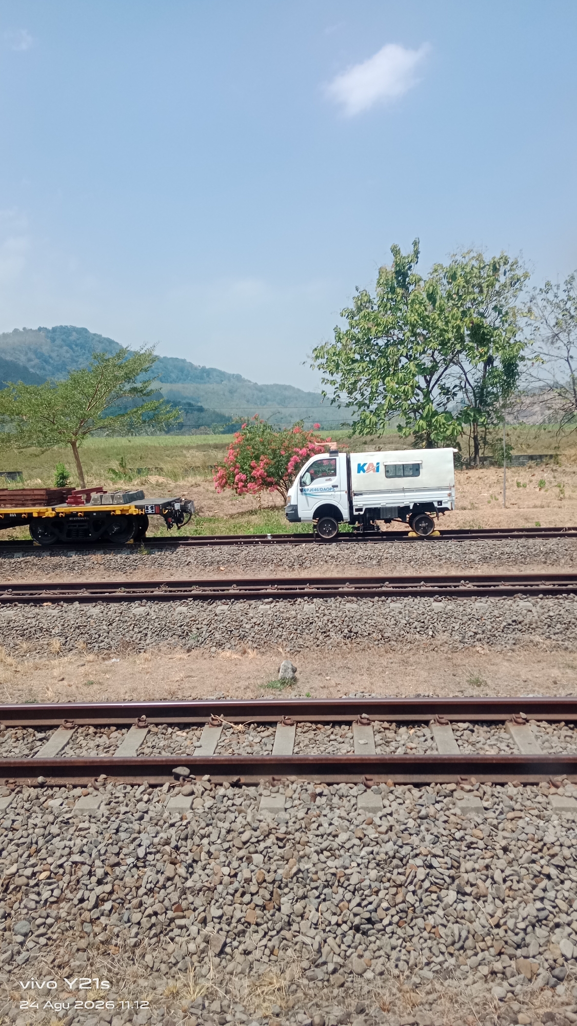 A white utility truck with "KAI" logo and license plate "APJC46/DAOP3" is parked on a railway track. The truck has a white cargo box with windows. A flatbed train car with some wooden planks is parked on an adjacent track. In the background, there are trees, some with pink flowers, and a hilly landscape. The scene is set during the day with a clear blue sky. The location is identified as Prupuk Utara, Indonesia, with the date and time stamp "24 Agu 2026 11:12" and device "vivo Y21s" in the lower left corner.
