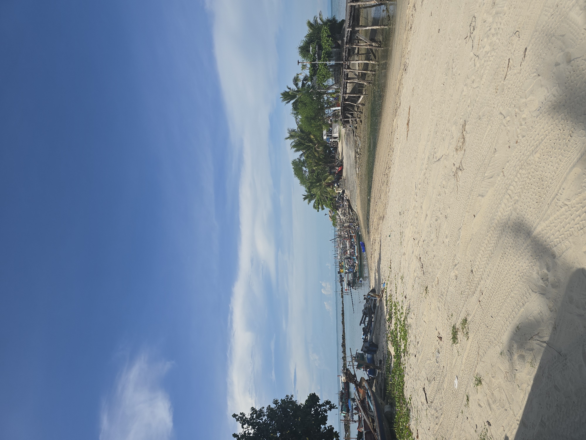 The image depicts a coastal scene in Baan Chaweng Noi, Thailand, under a clear blue sky with scattered white clouds. The foreground is dominated by a sandy beach with tire tracks and scattered debris. To the left, a large tree with lush green foliage stands near the water's edge.
The middle ground features a calm body of water where numerous small fishing boats are anchored. Some boats are pulled up onto the sand. Further out, a small island or peninsula is visible, densely covered with palm trees and other vegetation. A weathered wooden pier extends from the land towards the water, connecting to buildings on the island.
The background shows the sea meeting the horizon under the expansive sky. The scene appears peaceful and suggests a typical day at a fishing village. No people are clearly visible in the image.