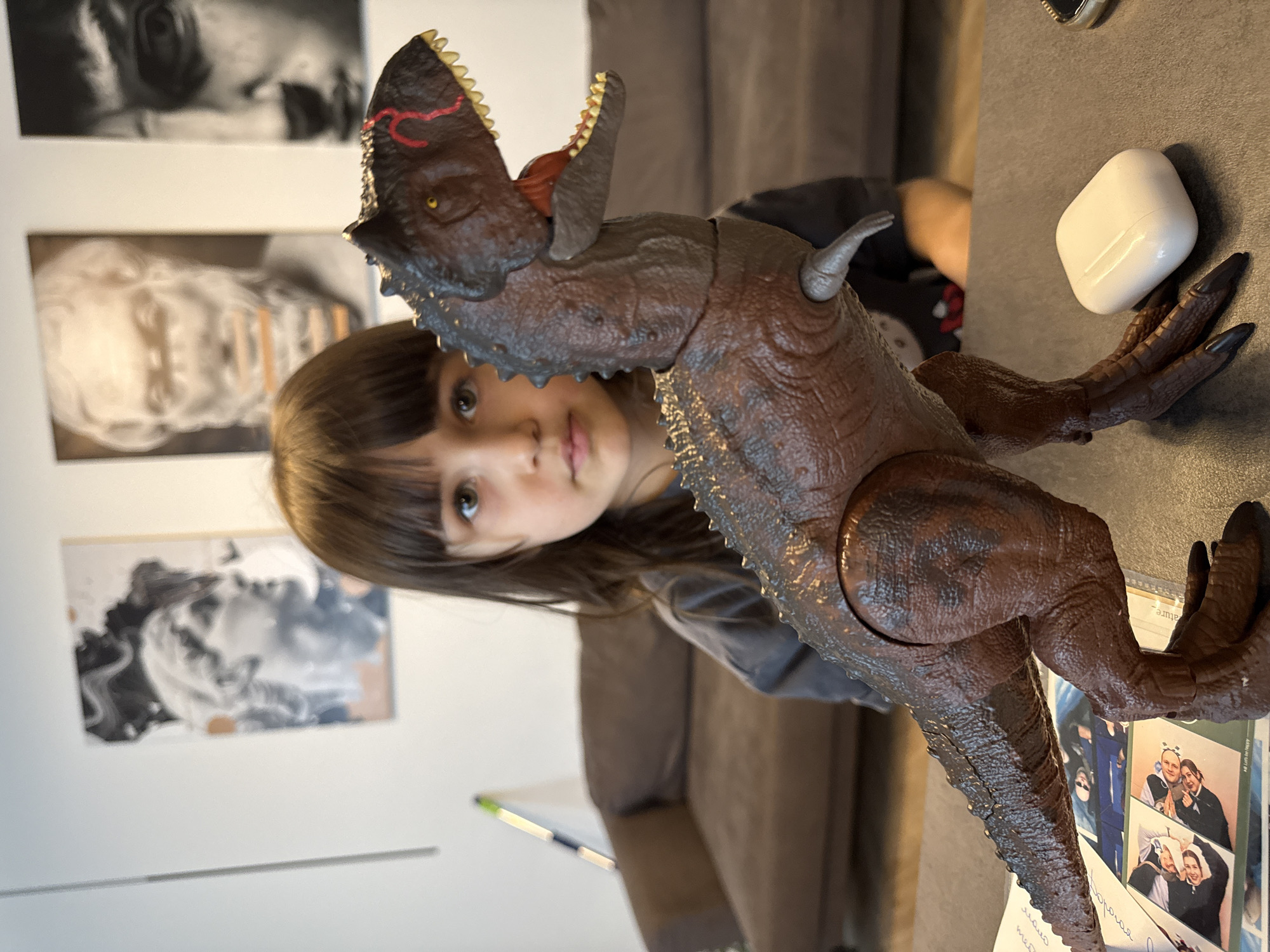 A young person stands behind a brown dinosaur toy with an open mouth, sharp teeth, and raised arms. The dinosaur appears to be holding a white object, possibly an electronic device. The scene is indoors, with artwork hanging on the wall behind the young person and a sofa in the background. There are photographs under the legs of the dinosaur. It is not possible to determine the exact location or city from this image.