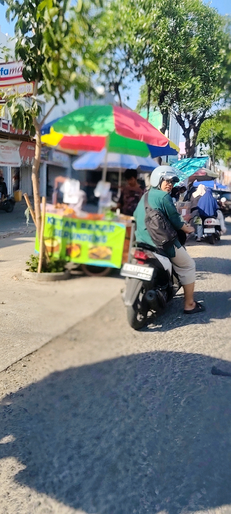 In the city of Cibatu, Indonesia, on a sunny day, a person wearing a teal shirt and light-colored pants rides a black motorcycle. They are also wearing a light blue helmet and a dark sling bag across their chest. The motorcycle has a license plate that reads "B 4112 KAD". The rider is looking towards the left of the frame. In the background, there is a food stall with a colorful umbrella, and a sign written in Indonesian. The food stall appears to be selling some sort of fried food. There are other people and motorcycles visible in the background, suggesting a busy street. The environment is somewhat blurry, indicating the image might have been taken while in motion.
