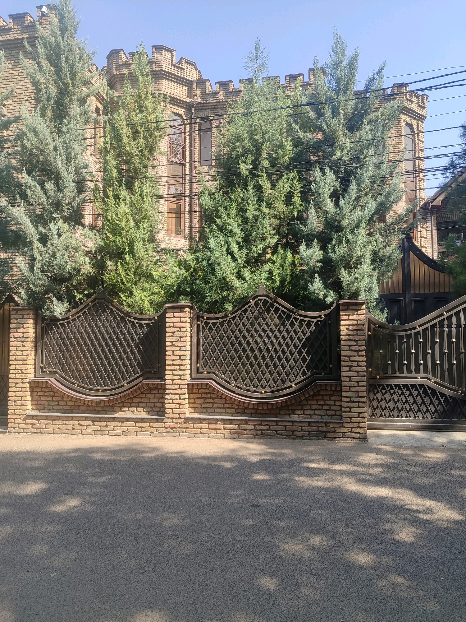 The image shows a brick building with castle-like crenellations and large windows, surrounded by numerous evergreen trees. A decorative metal fence with a brick base runs in front of the building. A street with dappled sunlight and shadows is visible in the foreground. The scene appears to be a residential area, likely a private residence, in Tashkent, Uzbekistan. There are no people or animals visible in the image. The sky is clear and blue.