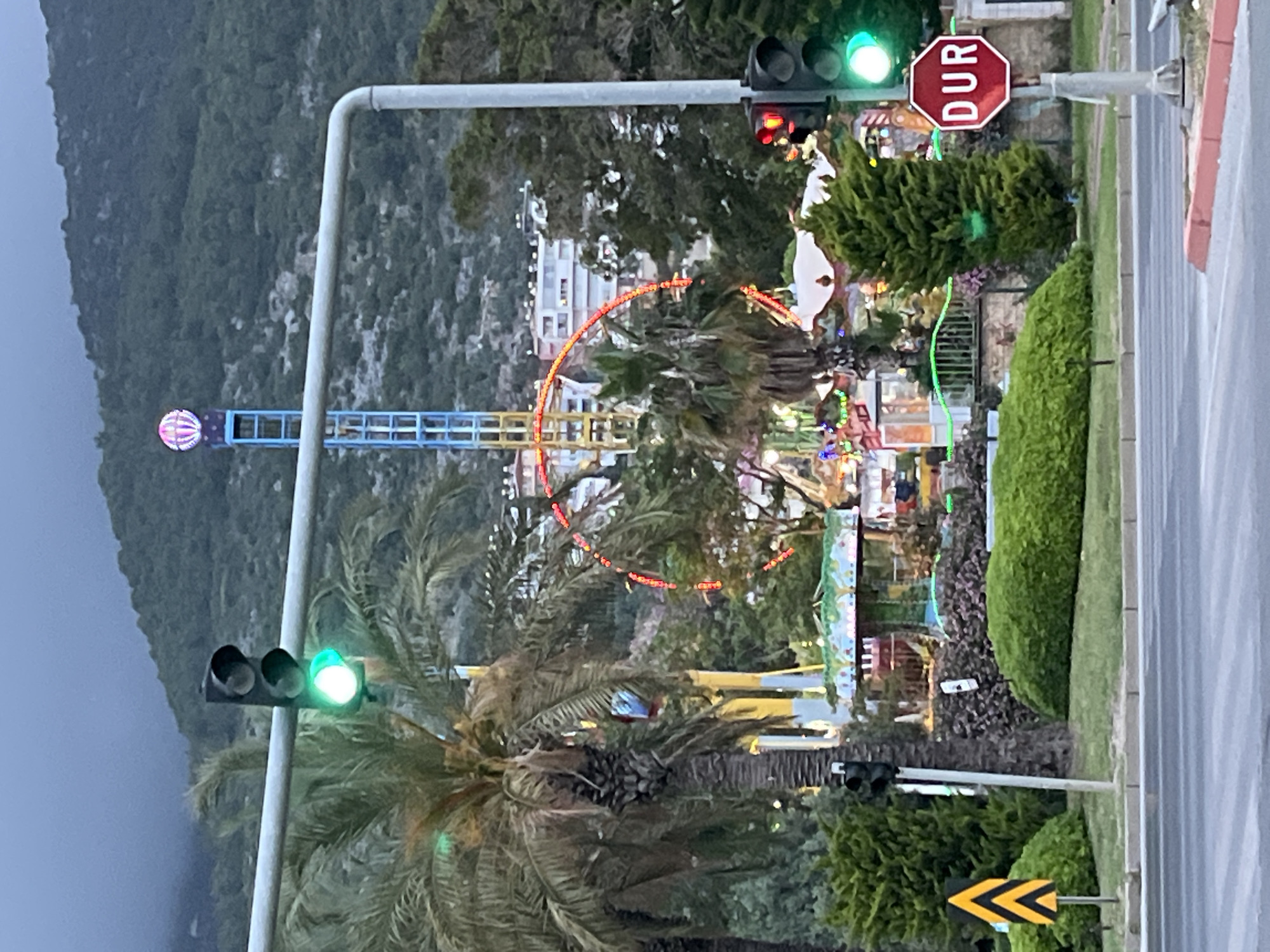 The image shows a view of a small amusement park in Alanya, Turkey, partially obscured by trees and traffic signals. In the background is a Ferris wheel with red lights, and a tall ride with a pink and white striped ball at the top. A green traffic light is visible in the foreground on both sides of the road. On the right side of the road is a red "DUR" (Turkish for "Stop") sign, and a yellow and black chevron road sign. There are buildings and a mountain in the background. The scene appears to be taken from a slightly elevated position.