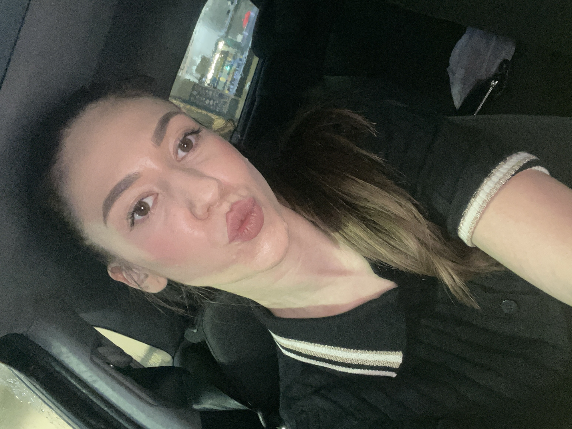 An indoor, close-up selfie captures a person, likely female, inside what appears to be a car, with the general location being Sofia, Bulgaria. The individual is positioned towards the left of the frame, looking directly at the camera with a playful "kissy face" expression, puckering her lips. Her dark brown eyes are visible, along with well-defined eyebrows and subtle makeup, including blush on her cheeks. Her hair, a blend of brown and lighter highlights, is swept back and falls over her shoulder.
She is wearing a dark-colored top, possibly black, with a distinctive ribbed collar or trim featuring horizontal stripes in white, beige/gold, and black. The car's interior is largely dark, with a dark headliner visible above. Through the side window in the upper right, blurred and diffused artificial lights suggest an urban environment at night. The window surface appears wet, indicating rain or condensation, which further blurs the exterior scene. In the background, behind the person's right shoulder, a small, crumpled white object, possibly a tissue, is seen near a dark zipper pull. The overall mood conveyed is lighthearted and personal. No visible text is present in the image.