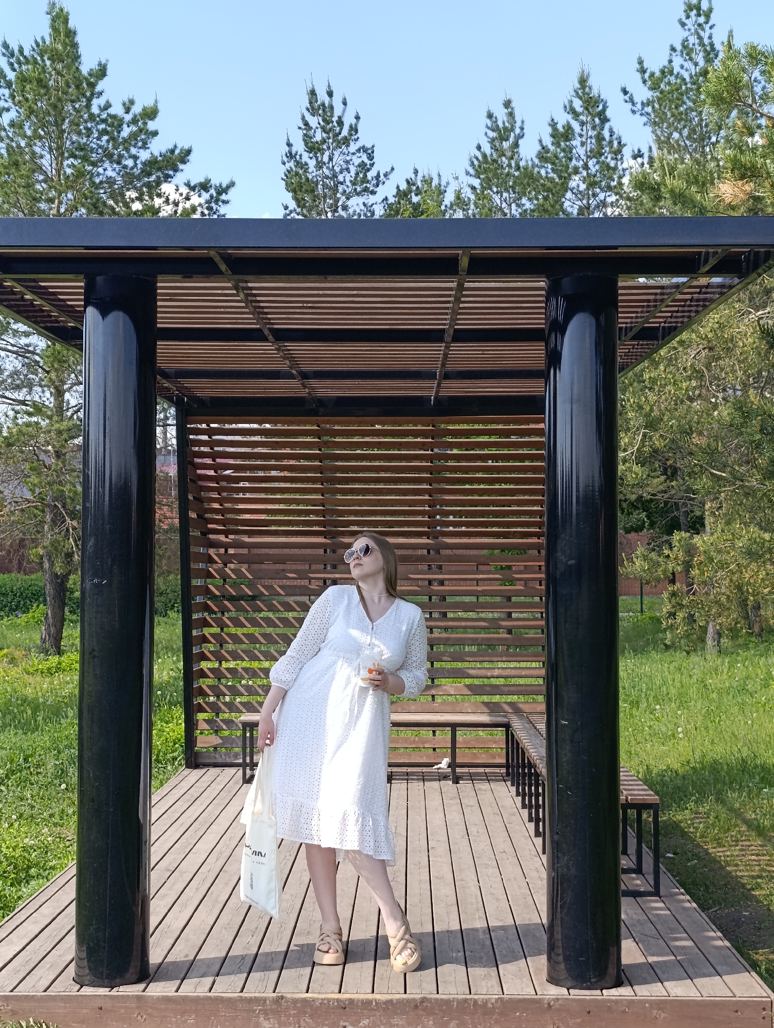 A young woman in a white dress stands in a wooden pavilion. The pavilion has a grid-like wooden wall and a wooden floor, supported by four black cylindrical posts. The woman holds a white tote bag in her left hand and a beverage in her right hand. She wears sunglasses and beige platform sandals. The pavilion is situated in a grassy area with trees in the background. The sky is visible above the trees.