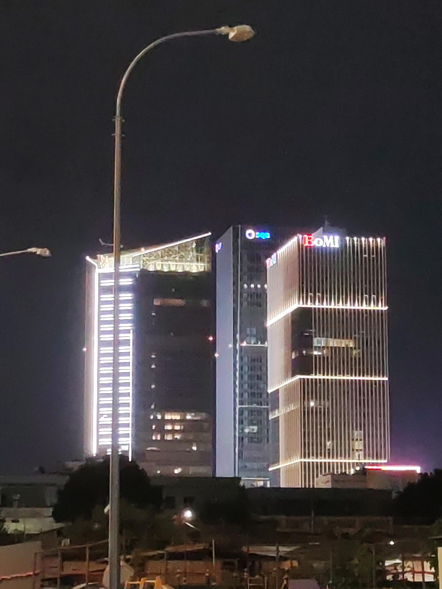 This is a night-time photograph of the Tashkent City urban development project in Tashkent, Uzbekistan. The image features several modern skyscrapers illuminated by an array of lights. In the foreground, a street lamp with a curved arm extends from the left side of the frame, obscuring a portion of the buildings. Two prominent buildings are visible: a taller, narrower skyscraper on the left with vertical white lighting strips, and a wider building to its right adorned with vertical golden lights and horizontal bands of light. The rightmost building displays the illuminated sign "BoMI" in red and white letters. Another smaller, blue illuminated sign, possibly "O'zbekiston", is visible on the left skyscraper. The background is dark, indicating nighttime, with the cityscape of Tashkent City illuminated against the night sky. In the lower part of the image, the silhouettes of trees and some construction elements are visible, suggesting ongoing development in the area.
