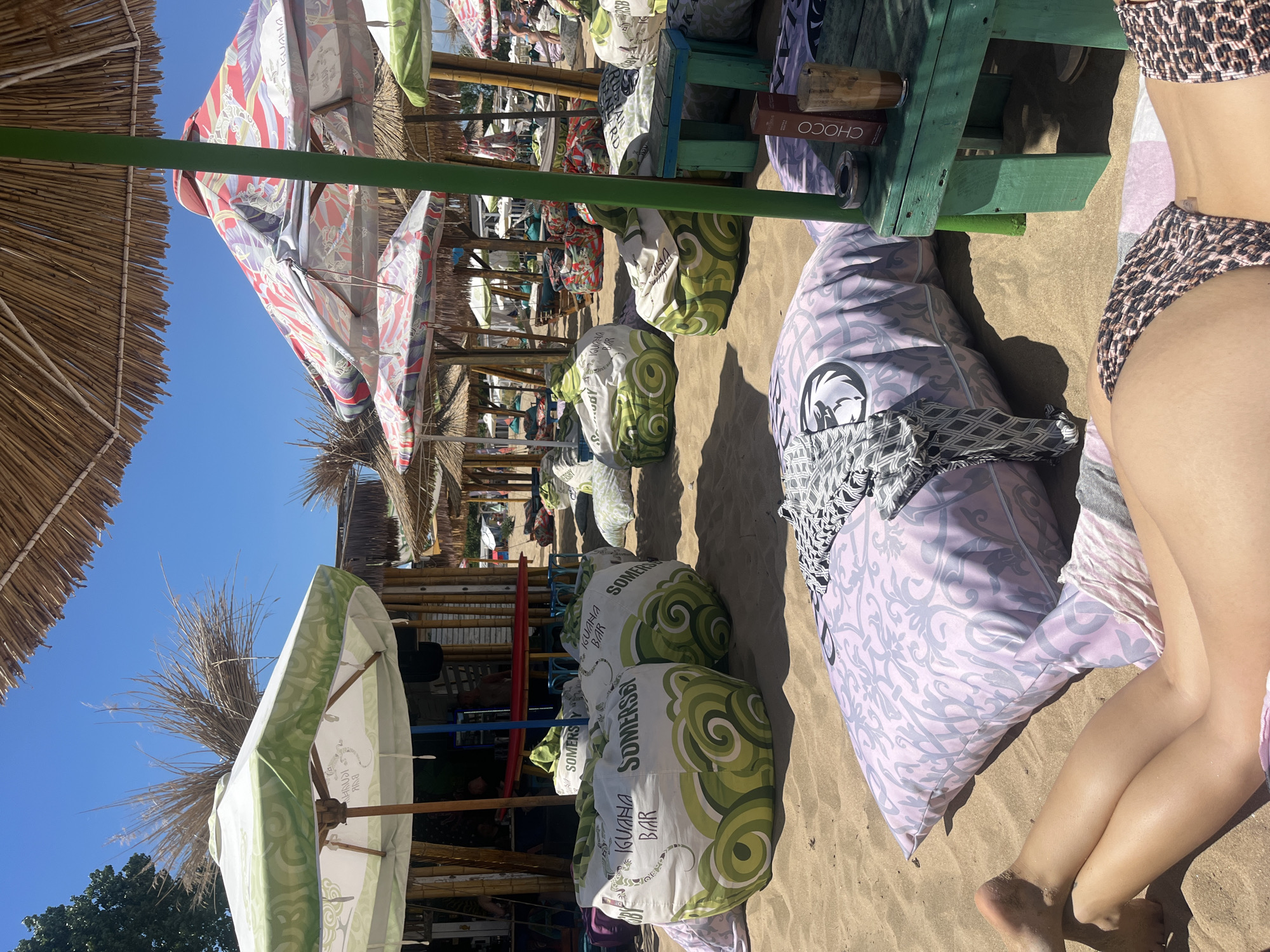 This image depicts a beach scene in Sozopol, Bulgaria, characterized by a vibrant beach bar and lounge area. In the foreground, a person lies on a large, patterned pillow, their legs visible in a leopard-print bikini bottom. To the left of the pillow, another person's legs are also visible, wearing striped shorts. The beach is sandy, with numerous large beanbag chairs scattered around.
Dominating the mid-ground are several of these beanbag chairs, some white with green circular patterns and the text "SOMERSET IGUANA BAR," others with different colorful patterns. Behind these beanbags, a bamboo-constructed beach bar is visible, with patrons seated inside. Several umbrellas, some with thatched roofs and others with colorful fabric, provide shade over the seating areas. A small, distressed green table stands to the right, holding a glass with a dark beverage and a box labeled "CHOCO."
The background shows more beach umbrellas and seating, with a clear blue sky overhead. The overall atmosphere is relaxed and summery, suggesting a typical day at a beach resort.