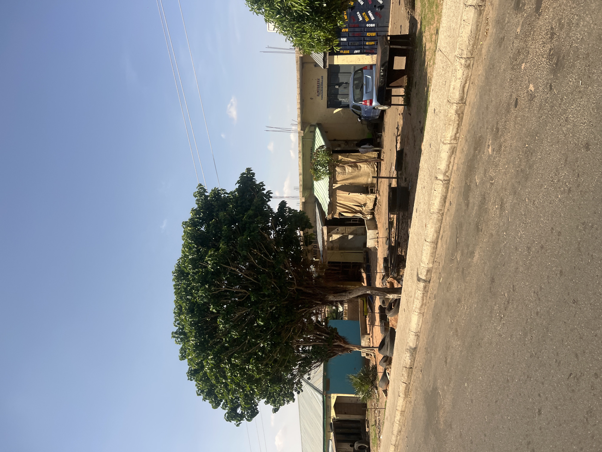 This image depicts a roadside scene in Gombe, Nigeria, under a bright, clear blue sky with minimal cloud cover and overhead utility lines traversing the upper right quadrant. The foreground features an asphalt road, appearing worn and soiled, bordered by a concrete curb separating it from an unpaved, reddish-brown earth area.
Along this unpaved strip, several single-story structures are visible. On the left, a building with a light blue corrugated metal roof and a light yellow facade transitions to a turquoise blue section. Towards the center and right, a light yellow/beige building with a partial flat roof, displaying exposed rebar, features a green corrugated metal awning over a covered outdoor section. A sign on this building reads "ALMUBAHAK FARHA ZAINAB ZAHRA ALI". To its right, a large dark blue metal gate is stenciled with various truck brand names including "IVECO," "DAF," "HOWO," "MACK," and "MAN."
Numerous discarded vehicle tires are scattered across the unpaved ground, particularly in the left and central areas. A large, mature tree with dense green foliage and visible trunks stands prominently on the left, with smaller trees and bushes nearby. Another tree is partially visible on the far right. A light blue compact car is parked in front of the central building. In the mid-ground, a dark, heavy-looking outdoor table or workbench is positioned on the dirt. One individual is partially visible, facing away from the camera, near the covered outdoor section of the central building. The overall scene suggests an active, possibly automotive-related, commercial or workshop area.