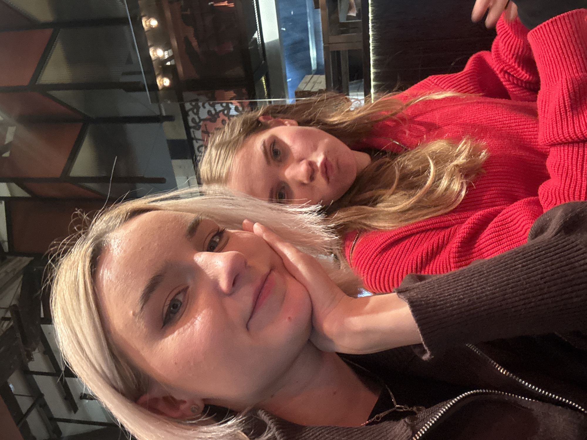 Two young women are posing for a selfie indoors. The woman in the foreground has short, light blonde hair and is wearing a dark jacket. She is touching her cheek with her hand. The woman in the background has long, blonde hair and is wearing a red sweater. The location is likely a restaurant or bar, based on the background with lighting fixtures and metal structures. The city and specific location in Russia are unknown.