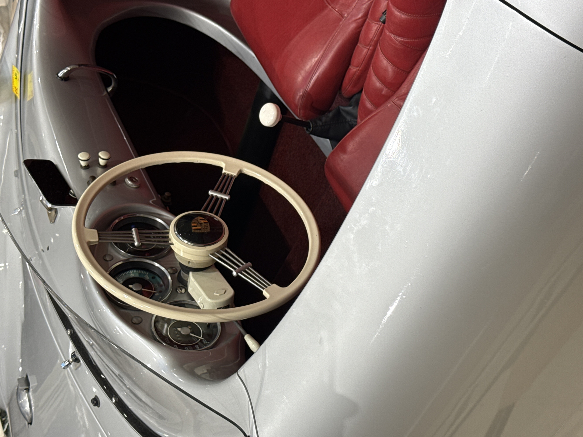 The image presents the interior of a classic silver Porsche, focusing on the dashboard and seating area. A cream-colored steering wheel with a Porsche emblem takes center stage, positioned in front of gauges and controls. Red leather seats are visible in the background. The car appears to be indoors, possibly at a car show or museum. The location is not explicitly specified as being in Rome, Italy.