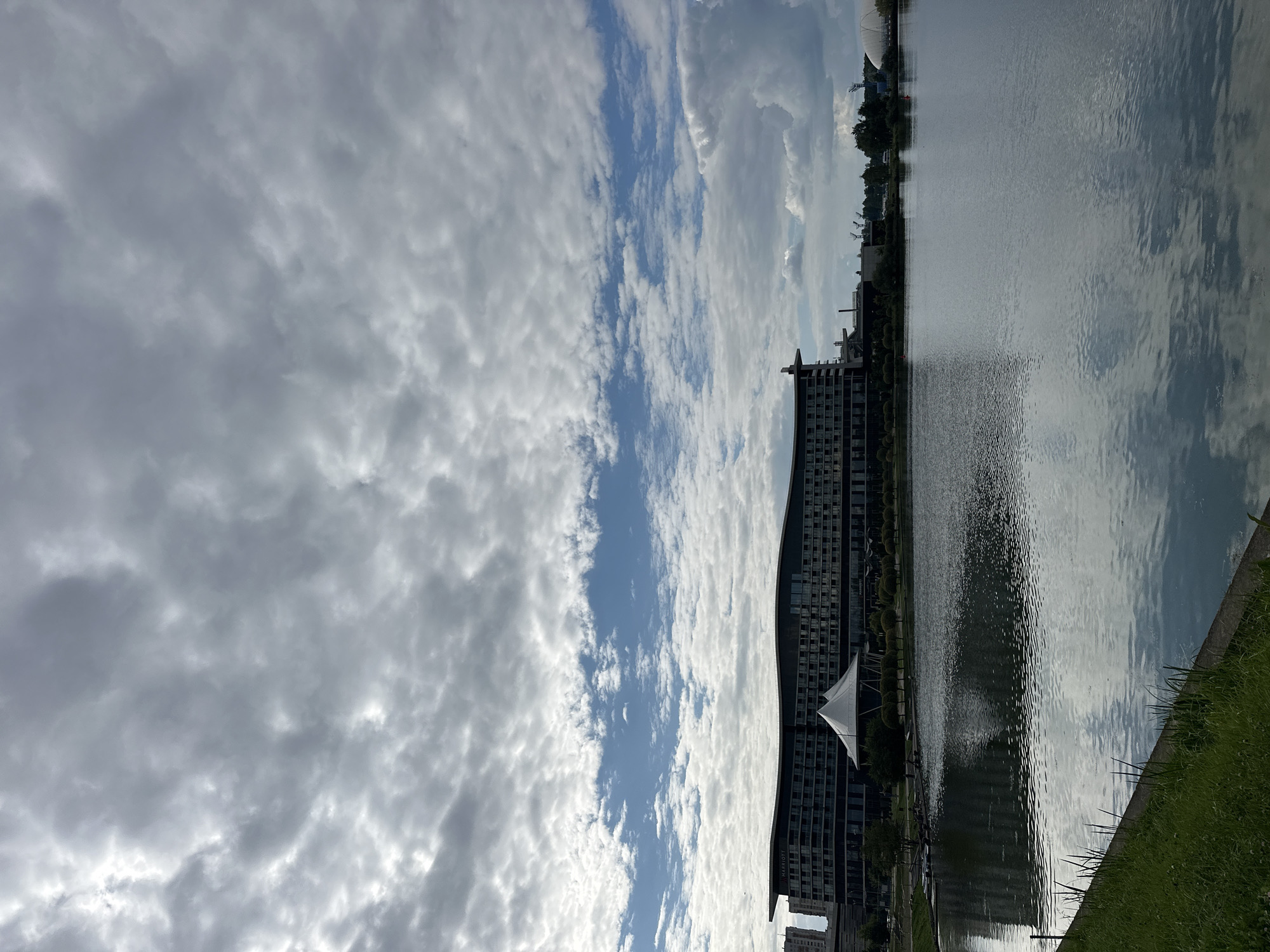 The image shows a large, modern hotel with a curved roof and many windows, labeled "MARRIOTT," situated on the bank of a calm lake. In front of the hotel, there is a white, tent-like structure. The lake's surface reflects the sky and the hotel. The foreground features a grassy bank with a concrete edge. The sky is filled with a mix of dark and light clouds, with patches of blue sky visible. In the background, trees and other buildings can be seen, including a domed structure on the right. The overall scene suggests an urban waterfront area. This location appears to be the Marriott Hotel in Minsk, Belarus, situated next to the Svislach River or a similar body of water.