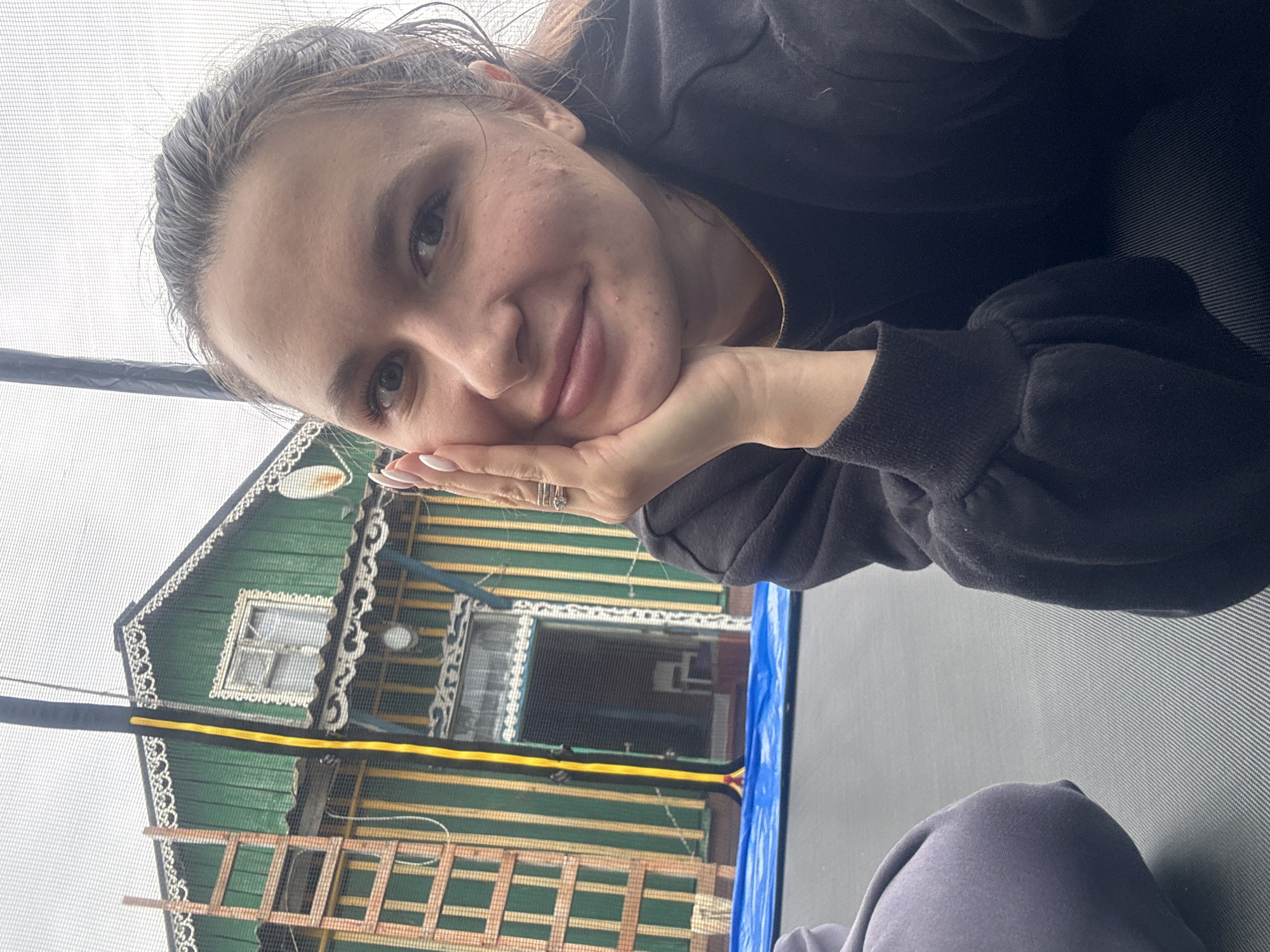 A woman is lying on a trampoline, resting her head on her hand and smiling at the camera. She is wearing a dark sweatshirt. Behind her is a two-story wooden building with green and yellow stripes and white decorative trim. A wooden ladder is leaning against the building. The image was taken in Красный Маяк, Russia.