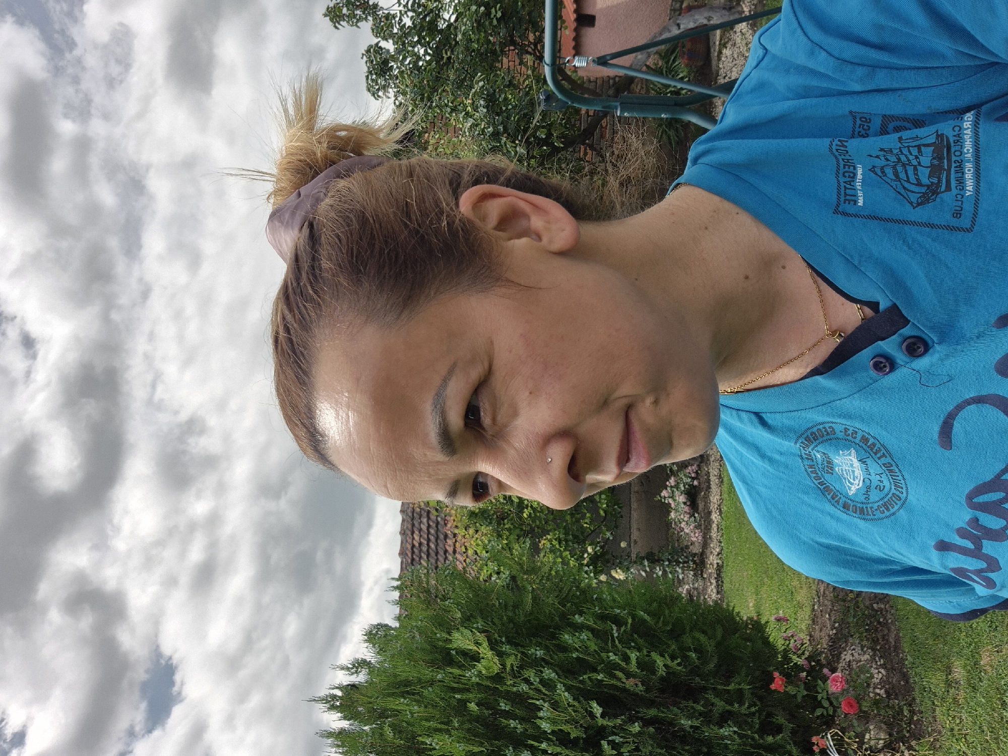 The image is a close-up, profile view of a woman in a backyard. The woman has blonde hair pulled back in a ponytail and is wearing a bright blue t-shirt. She has a nose ring and a gold chain necklace. Behind her, a green evergreen tree is prominent on the left side of the frame. Further in the background, a house with a tile roof and some greenery can be seen. The sky above is overcast with gray clouds. There is a swing set on the right side of the image, partially obscured by trees. The overall impression is a casual outdoor setting.