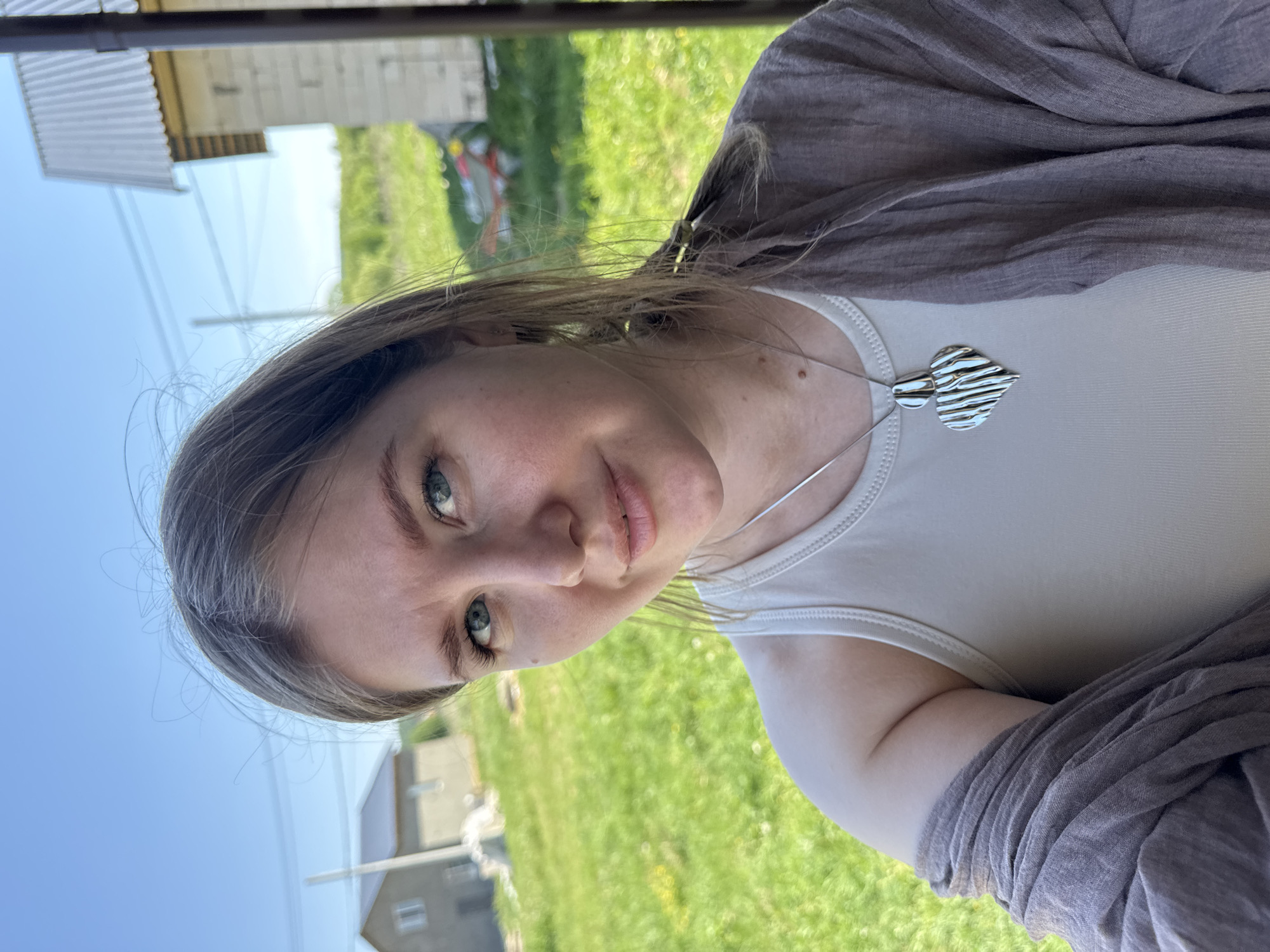 A young woman with light skin and light brown hair is centered in this outdoor selfie. She has blue eyes, light pink lips, and is wearing a light-colored tank top with a silver necklace. A brown shawl is draped over her shoulders. The background shows a green lawn and a few buildings, with one appearing to be a house. The location appears to be in a rural area. The selfie was taken outdoors in the city of Ферма, Russia.