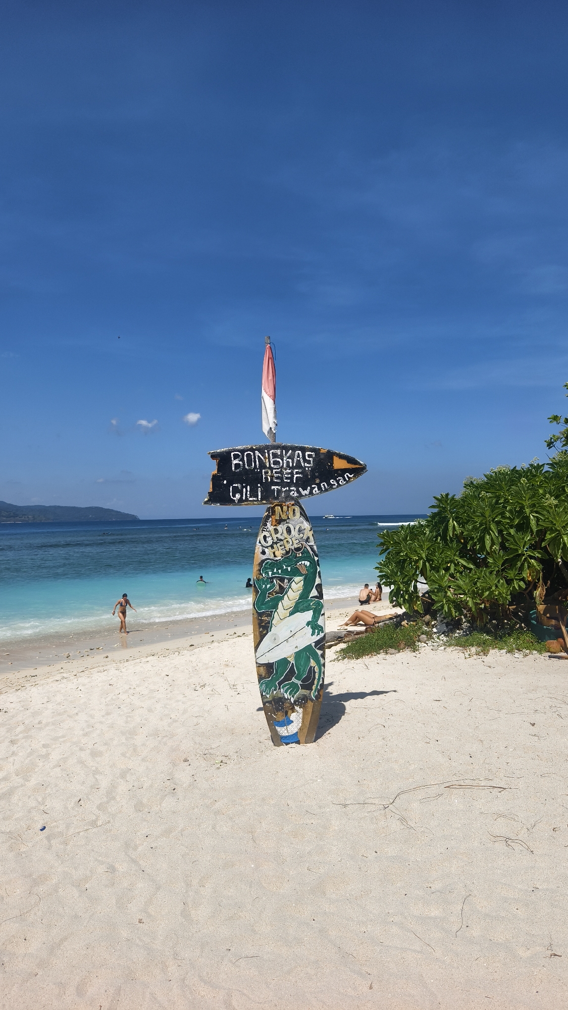 The image depicts a tropical beach scene in an unknown location in Indonesia. The sky is bright blue with a few scattered clouds. A signpost stands prominently in the foreground, made of an old surfboard and a directional sign. The directional sign reads "Bongkas Reef" and "Gili Trawangan", indicating the direction of these locations. The surfboard beneath has a cartoon crocodile painted on it with the text "NO CROC HERE". At the top of the sign is the Indonesian flag.
In the background, the turquoise ocean meets a sandy beach. A few people are visible; one person walks into the water, another is swimming, and two are sunbathing on the beach. Lush green vegetation is present on the right side of the frame. The sand appears light tan in color.