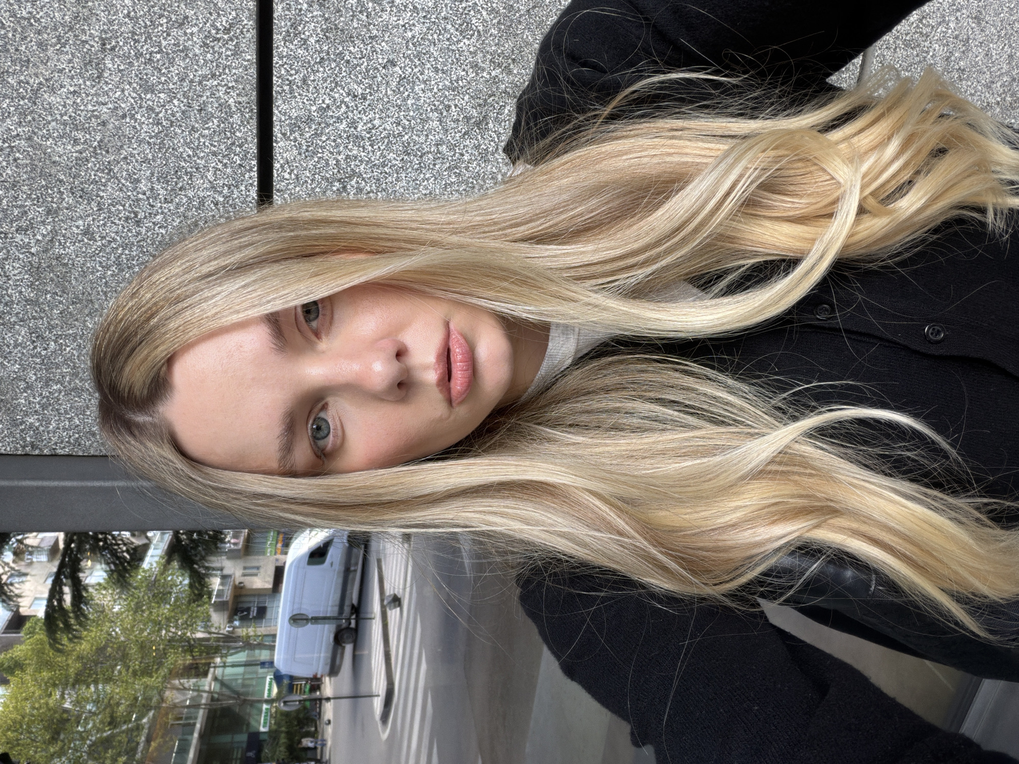 A close-up portrait of a young woman features her head positioned on the left side of the frame, looking directly towards the camera with light-colored eyes and a neutral expression. Her long, wavy blonde hair, with lighter highlights, flows out to the right, covering a significant portion of the image. She wears a black cardigan or jacket over a light-colored top. Her face shows light makeup, including lip gloss.
The background is split into two main sections. Behind her head and upper body is a textured, speckled grey wall, resembling granite or concrete, with a prominent dark vertical line running through it. In the lower right half of the image, reflecting on what appears to be a glossy surface (such as a car hood or an outdoor table), is a blurred urban street scene. This reflection reveals a white van, multi-story buildings, green trees, and a distinct zebra crossing on the road.
The image captures a relaxed moment in a bustling urban environment. It is a daytime shot, illuminated by soft, natural light, suggesting either morning or late afternoon. The location context for this urban setting is Tbilisi, Georgia. There is no visible text in the image.