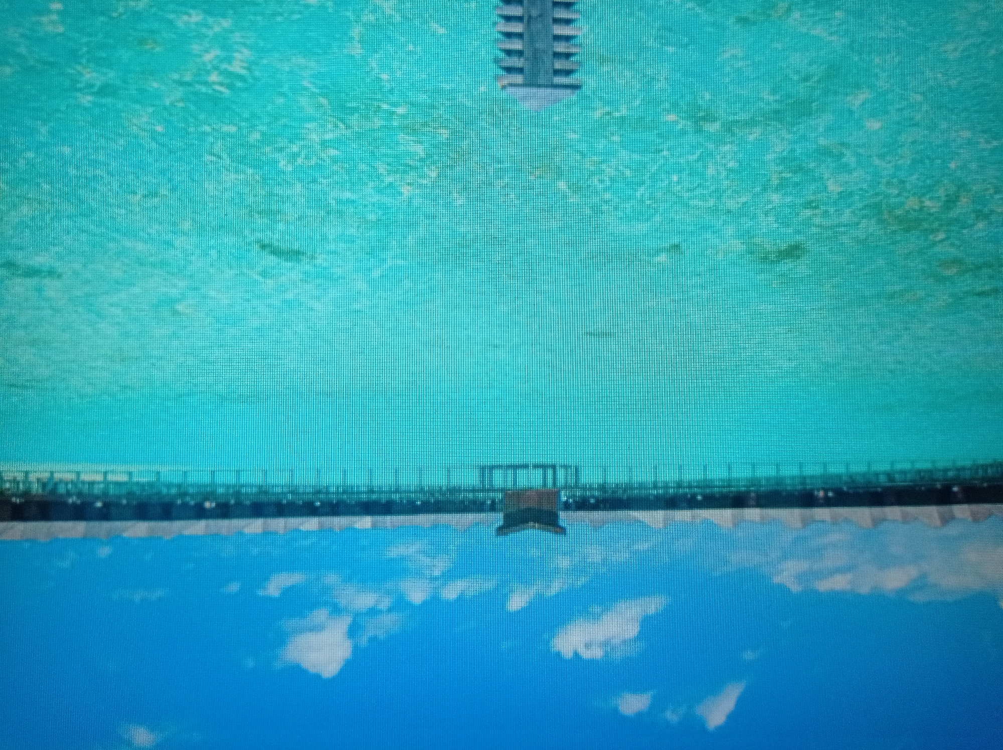 The image shows a picturesque view of a tropical resort with overwater bungalows. The turquoise water is clear, revealing the sandy bottom and some vegetation. A long wooden pier extends into the water, lined with numerous bungalows. In the center of the pier, there is a more elaborate structure with a dark, pitched roof. The sky is a vibrant blue with a few scattered white clouds. The scene is tranquil and idyllic, suggesting a vacation destination. The image is taken from a low vantage point, possibly from a boat or from the water itself, looking towards the resort. The foreground features the tip of what appears to be a light fixture or a piece of equipment, further suggesting the vantage point. The image is a photograph, and due to the resolution, it is difficult to discern specific details. This image is NOT from Mukachevo, Ukraine.
