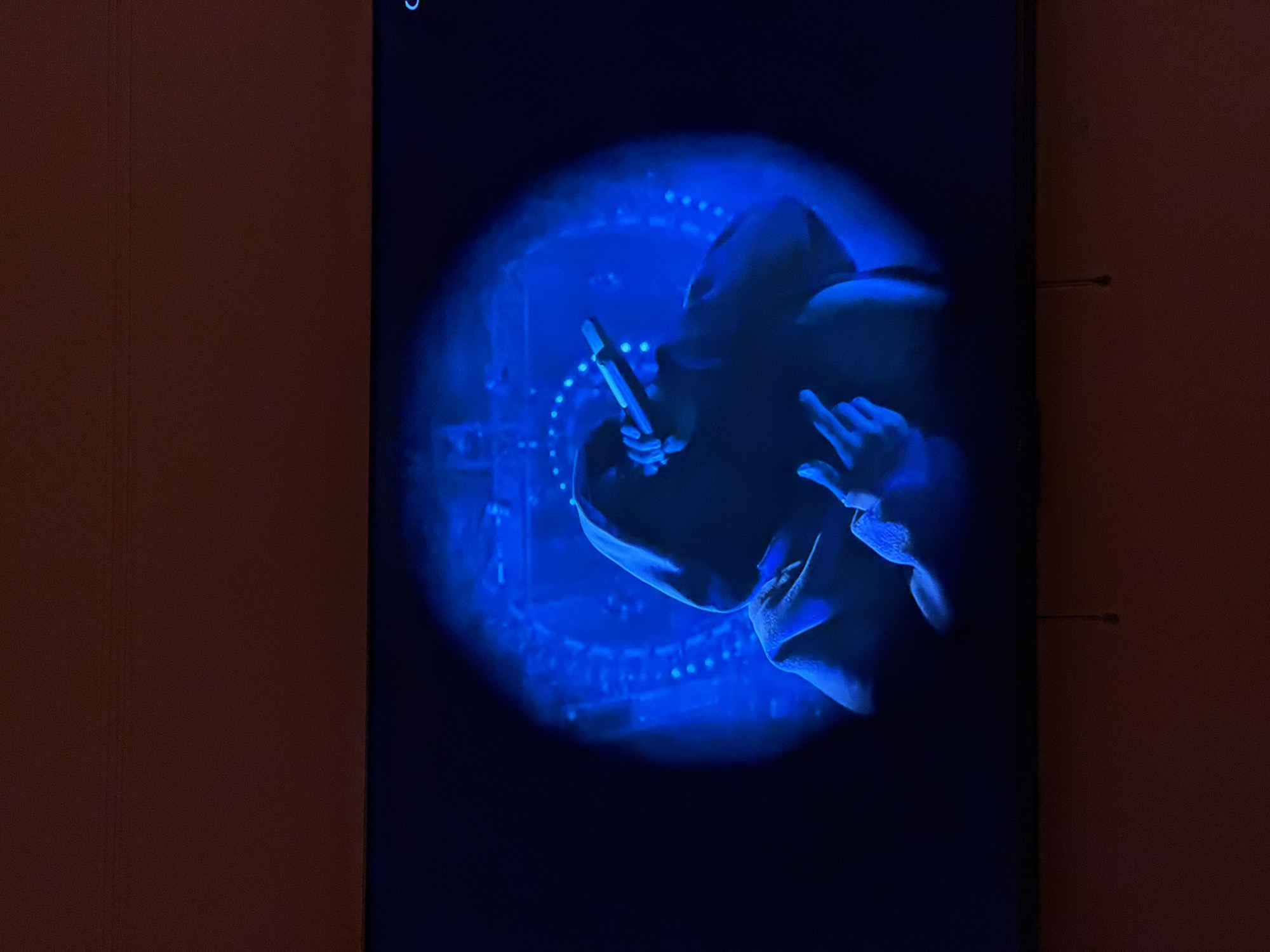 A close-up image of a person wearing a hooded sweatshirt is displayed on a screen, likely a television. The scene is illuminated by a blue spotlight, obscuring facial features and creating stark shadows. The person is holding what appears to be a microphone in one hand and making a hand gesture with the other. The background shows a stage-like setup with lights, suggesting a performance or event. The screen displaying the image is situated in a dimly lit room. Part of the wall is visible next to the screen. The location of the image is unspecified and can not be determined to be in the city of New York, United States of America.