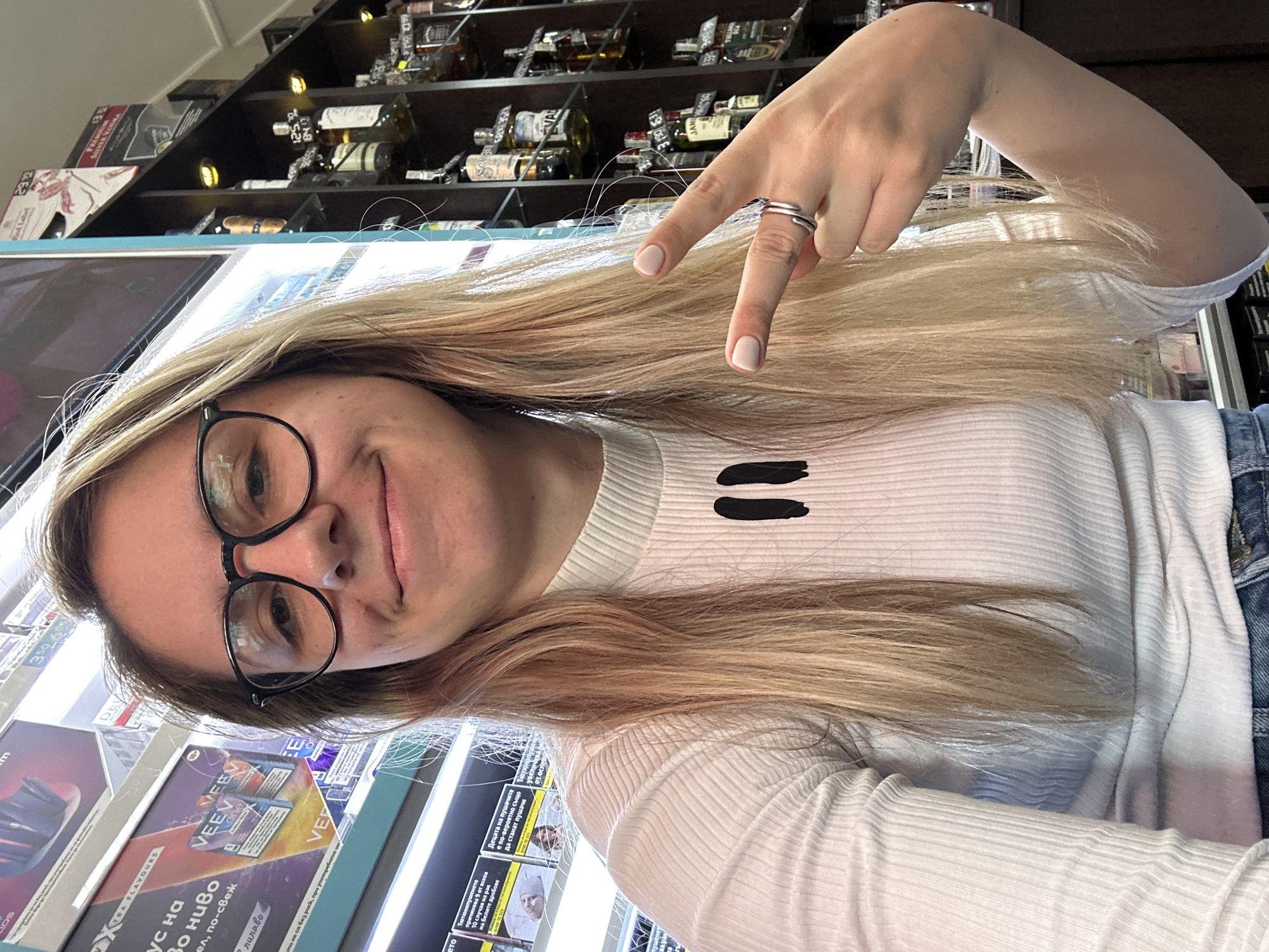 A young woman with blonde hair and glasses is taking a selfie in what appears to be a convenience store or a duty-free shop. She is wearing a white, ribbed, turtleneck shirt and blue jeans. Her left hand is raised, forming a peace sign, and she is wearing a ring on her left ring finger. Behind her, shelves are stocked with various products, including bottles of alcohol and what seem to be vape products. The background suggests an indoor commercial environment. The store appears to be located in Sofia, Bulgaria, based on the text visible on some of the product packaging.