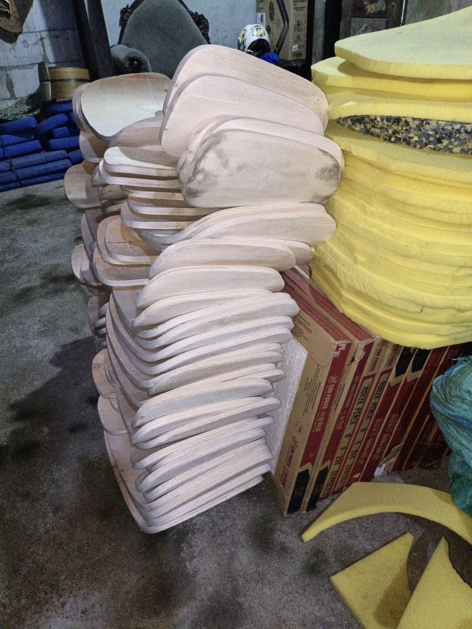 In Cikal, Indonesia, a dimly lit storage area is filled with stacked materials. The foreground is dominated by a large pile of light-colored, curved wooden pieces, possibly components for furniture or other manufactured items. To the right, a tall stack of bright yellow foam pads is visible. Between the wooden pieces and the foam, a cardboard box with red and white labeling, featuring Chinese characters and the text "SUN POWER," is present. In the background, stacks of blue rolled fabric or matting are seen next to a dark, upholstered chair. The floor is concrete, showing signs of wear and dust. The overall impression is of a workshop or storage facility awaiting further processing or assembly of these materials.