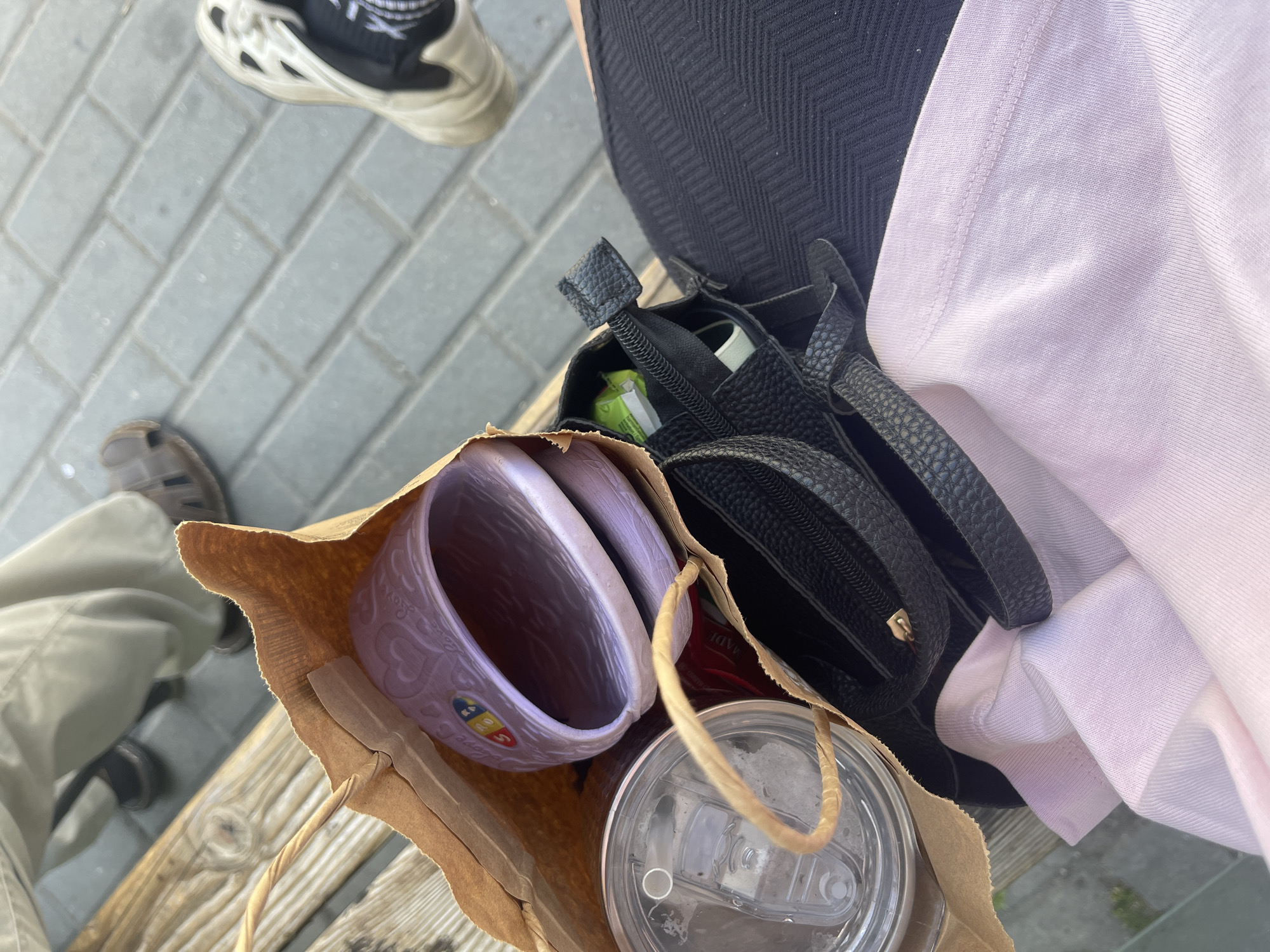 In Varna, Bulgaria, on a paved area adjacent to a wooden bench, a person sits with a brown paper bag and a black purse. The paper bag contains a translucent tumbler, a pair of light purple slides with heart patterns, and a red object, possibly a packet. A pink garment is draped over the purse. In the background, another person's legs are visible, wearing khaki pants and brown sandals, and another person's legs are visible wearing white sneakers. The lighting suggests it is daytime.