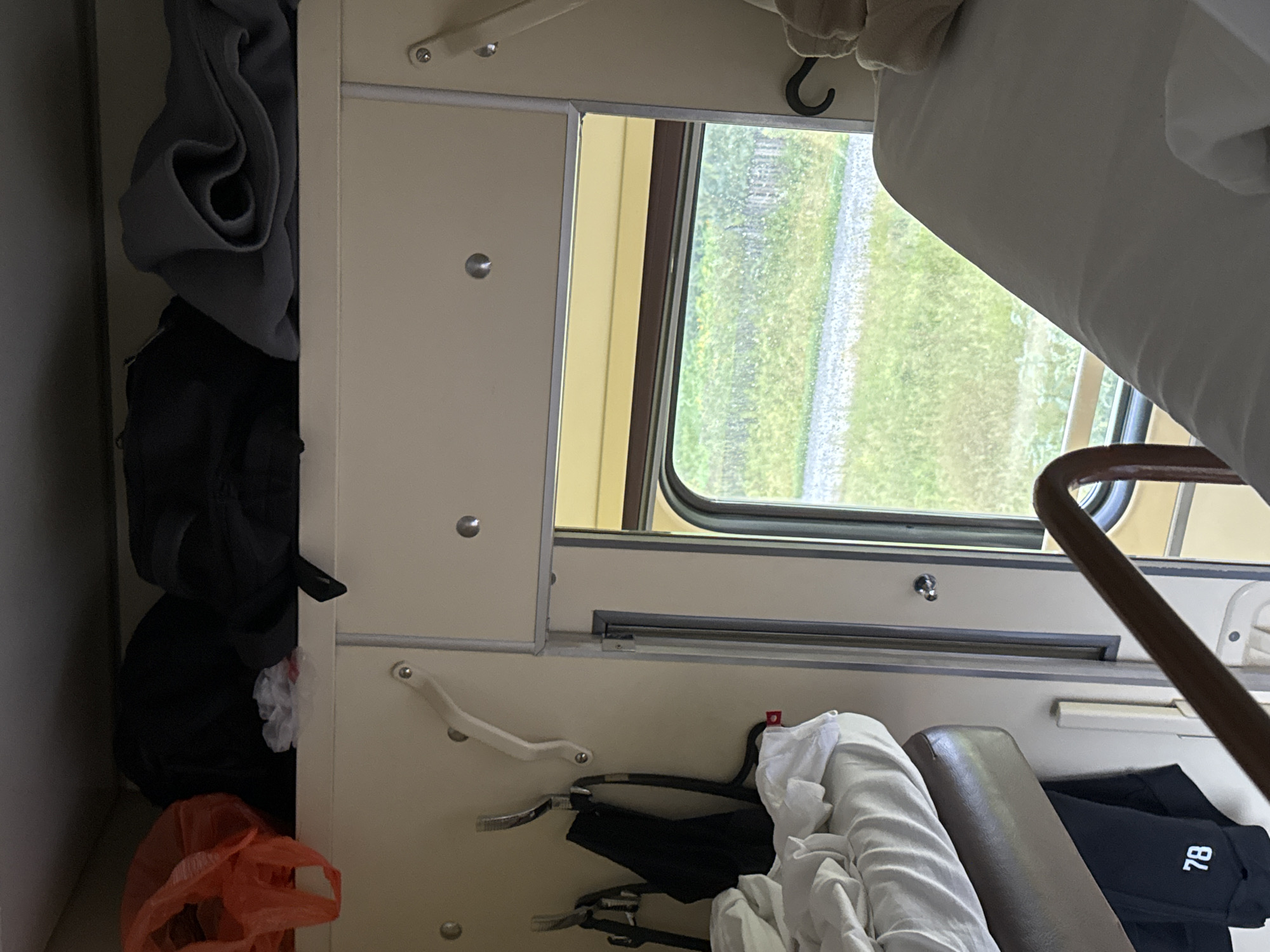 This image depicts the interior of a train compartment, likely in Shalya, Russia. The compartment contains two berths, one above the other. The upper berth is covered with white bedding, and a brown luggage rack is visible above it, holding several bags, including an orange plastic bag and two dark-colored backpacks. Below the upper berth, on the wall, are two metal hooks. A black garment, possibly pants, with the number "78" printed on it in white, hangs from a hook on the lower berth. Adjacent to the lower berth is a window looking out onto a landscape with green foliage and a gravel path. A metal handrail for the upper berth is visible in the foreground, and a portion of the lower berth is also visible. The walls and ceiling of the compartment are a light beige color. There are no people visible in the image.