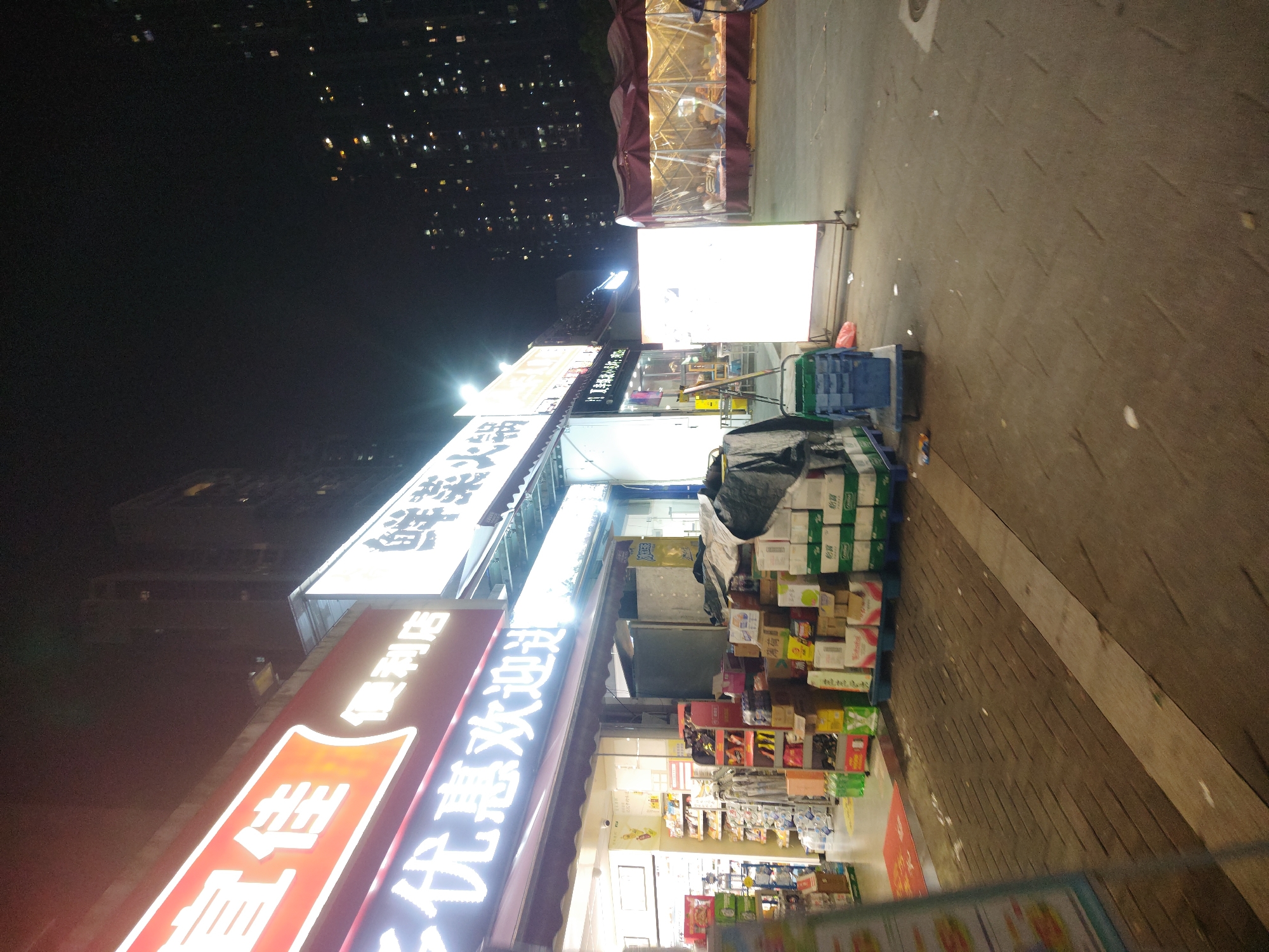 This image depicts a bustling urban street scene at night in the Chongqing High-tech Zone, China. The dark night sky is punctuated by numerous distant lights from high-rise buildings, indicating a densely populated city environment.
The foreground is dominated by brightly lit storefronts of what appear to be convenience stores and supermarkets. A prominent red sign with white Chinese characters clearly displays "直佳便利店" (Zhi Jia Convenience Store). Below this, a white illuminated sign reads "优惠欢迎" (Discounts Welcome). Further up, another large white sign designates "生活超市" (Life Supermarket).
Outside the convenience store, the sidewalk is stacked with numerous cardboard boxes and green/blue crates on a pallet, likely containing inventory for the store. A large, bright, freestanding light box also illuminates the area. Further down the street, a transparent, tent-like structure with a red frame is visible, possibly serving as a temporary outdoor stall or dining area. The ground is paved, and some minor litter is scattered. No distinct people are clearly visible, but faint figures might be present within the distant tent structure. The scene portrays typical nocturnal commercial activity in a Chinese urban setting.