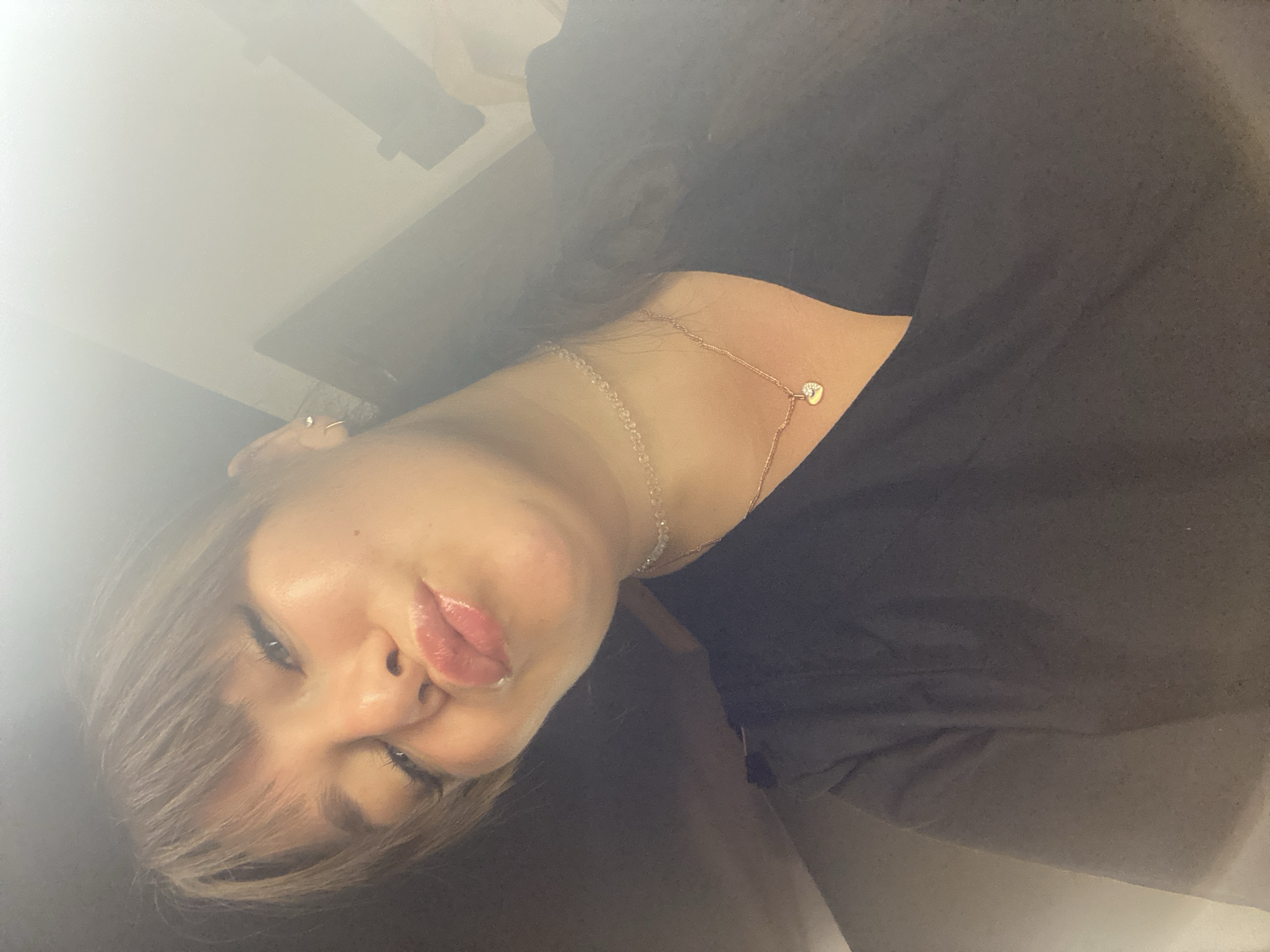 A young woman with bangs and a black V-neck shirt is captured in a selfie. She wears a delicate choker necklace and a rose gold chain with a heart-shaped pendant. Her lips are puckered, and she appears to be indoors, with a blurred background of what seems to be a wall and doorframe. The lighting is bright, creating a lens flare effect. There is no discernible information about the location in Votkinsk, Russia.