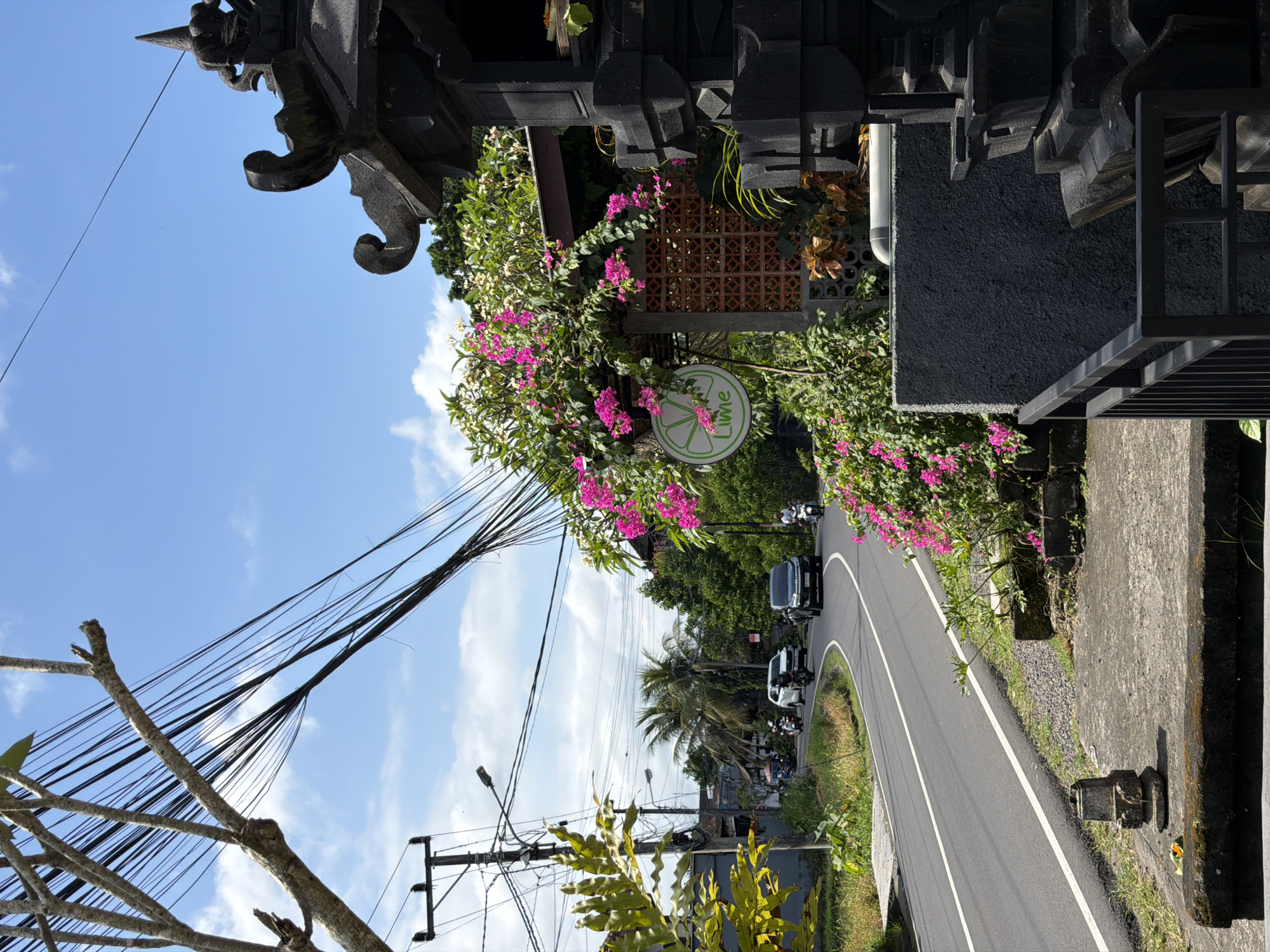 The image captures a street scene in Mengwi, Indonesia. In the foreground, an ornate, dark-colored structure frames the view, partially obscured by vibrant pink flowers and greenery. A round sign, marked with the word "Lime", is also visible amidst the foliage. A paved road curves into the distance, with two cars, a black SUV, and a white vehicle, driving along the road. Several scooters are also visible. The roadside is lined with vegetation, including palm trees and other tropical plants. Overhead, a web of power lines stretches across a blue sky dotted with clouds. A utility pole is also present on the side of the road.