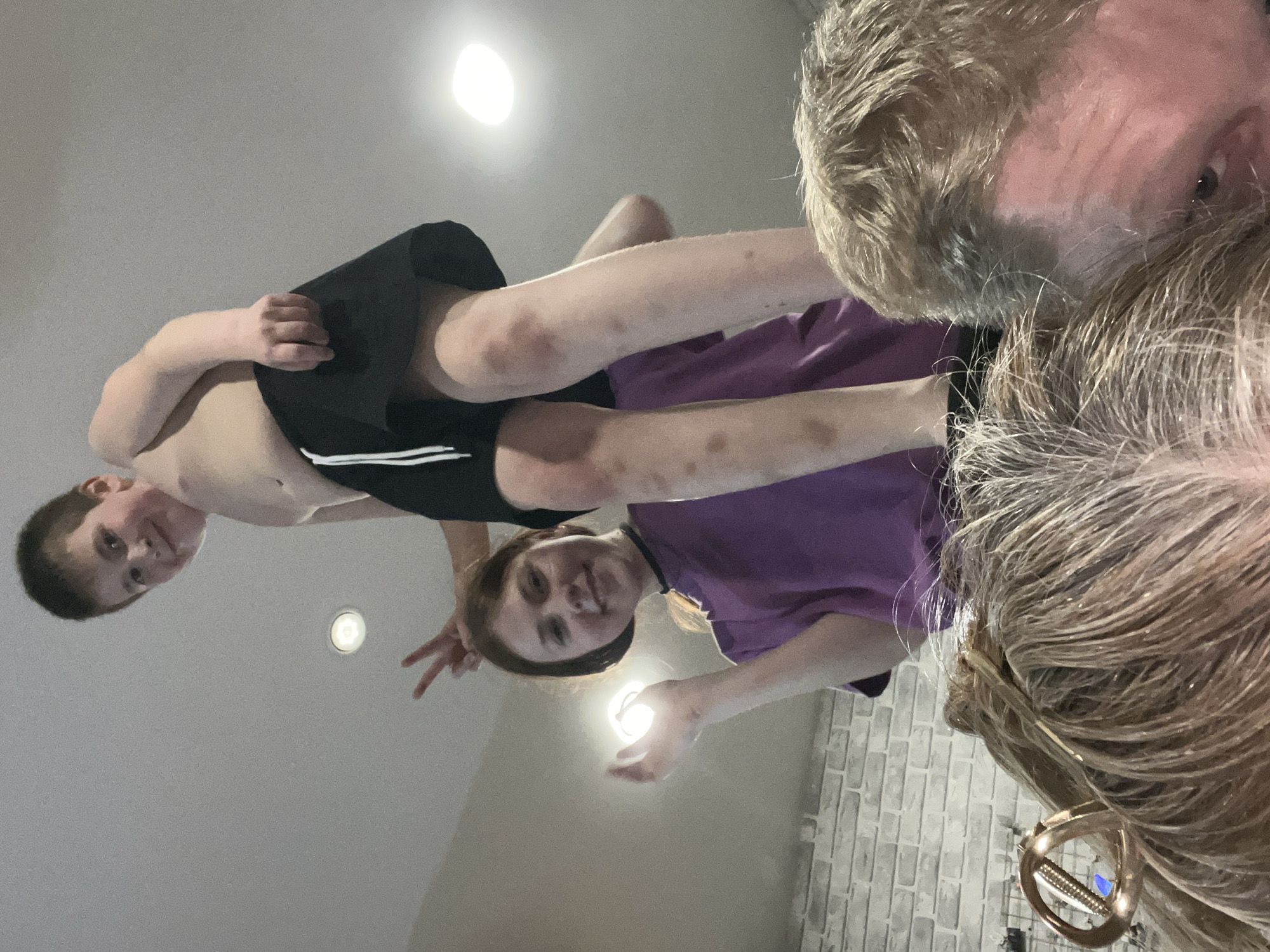 The photograph, taken from a low angle looking up, depicts four individuals. The subjects appear to be close to the camera, creating a foreshortened perspective. A boy in black shorts is positioned at the top left. A girl wearing a purple shirt and holding her fingers up in a "peace sign" can be seen at the lower left. At the right of the frame is an adult, with their face very close to the camera. The scene is indoors, illuminated by overhead lighting, as indicated by the light fixtures visible in the background. The environment includes a white ceiling and a brick wall. No information about Vesyoloye, Russia, is found.