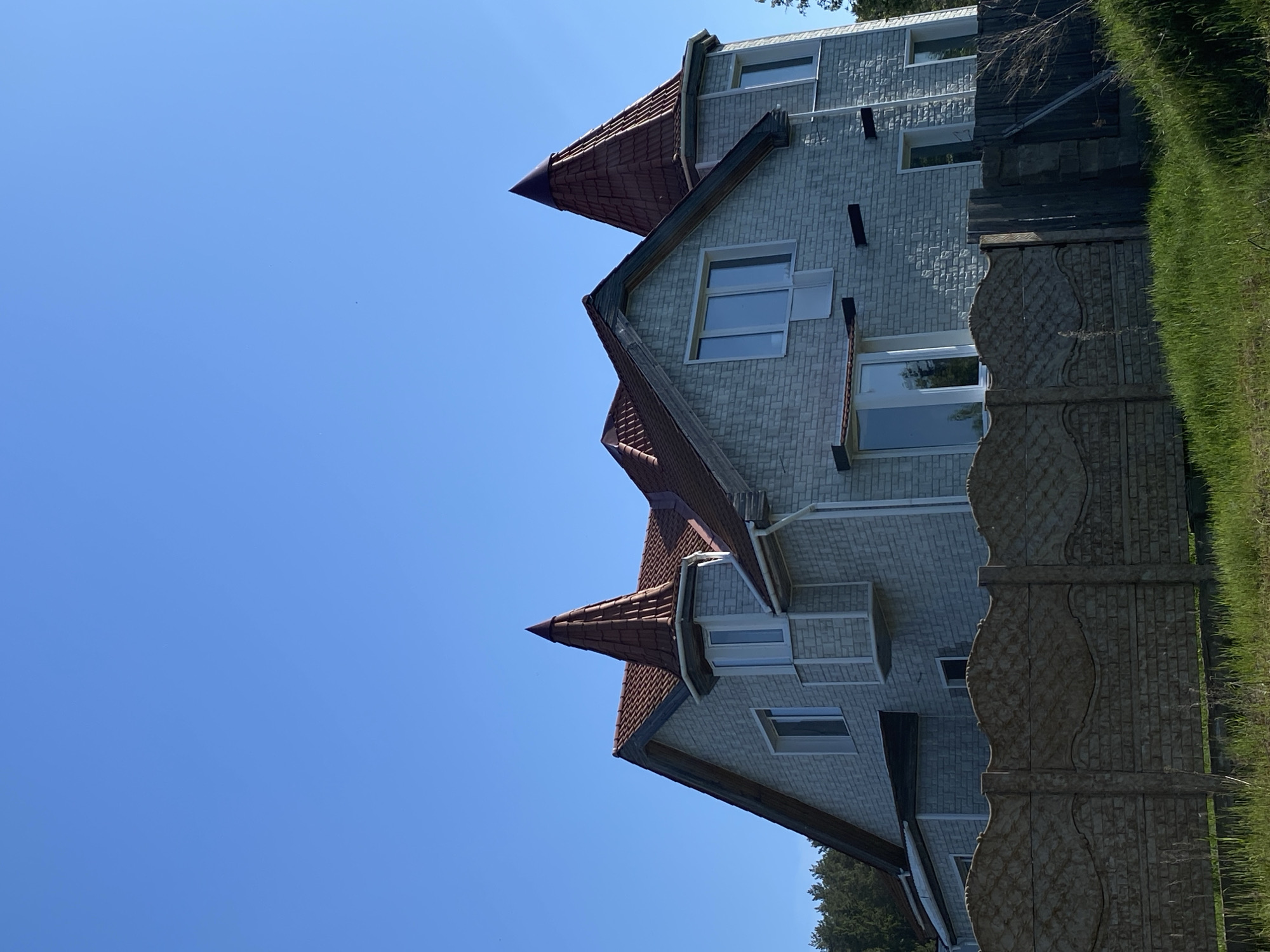 The image depicts a three-story building with multiple turrets topped with conical roofs. The building's facade is light gray brick with multiple windows. A decorative concrete fence runs along the right side of the frame, separating the building from a grassy area. The sky is a clear blue. The angle of the shot is tilted upward, capturing the building's height against the sky. It is not possible to independently confirm the location is in Votkinsk, Russia, based on the provided image.